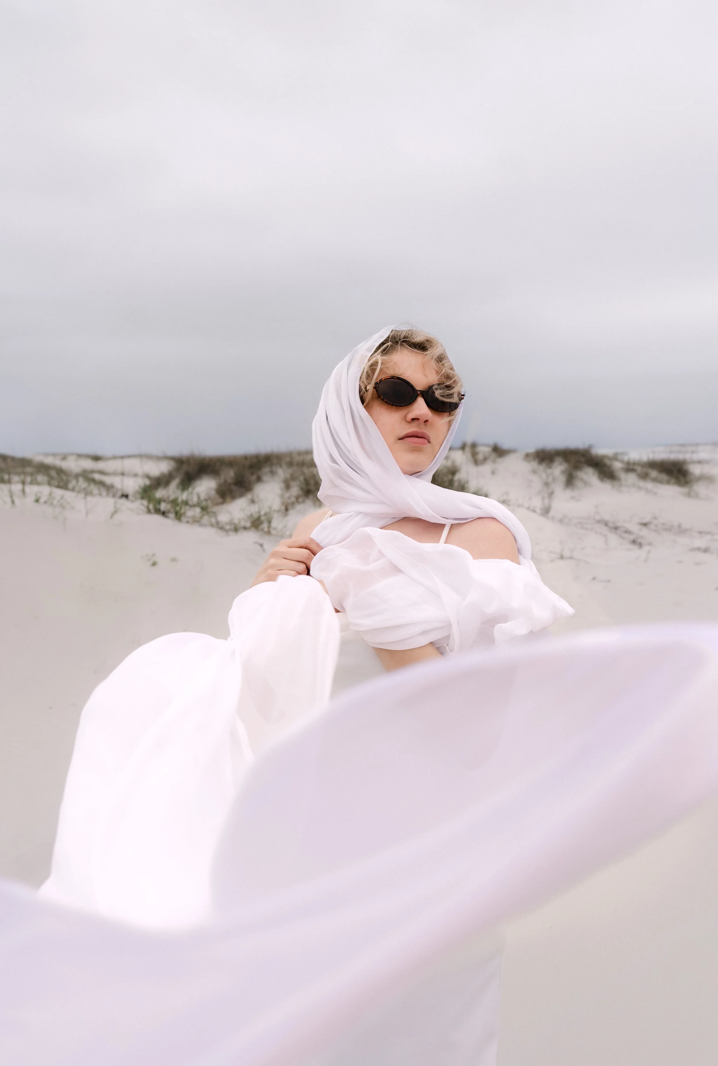 A woman with curly blonde hair wearing black sunglasses and a white headscarf, standing outdoors on a cloudy day with sand dunes and sparse vegetation in the background.