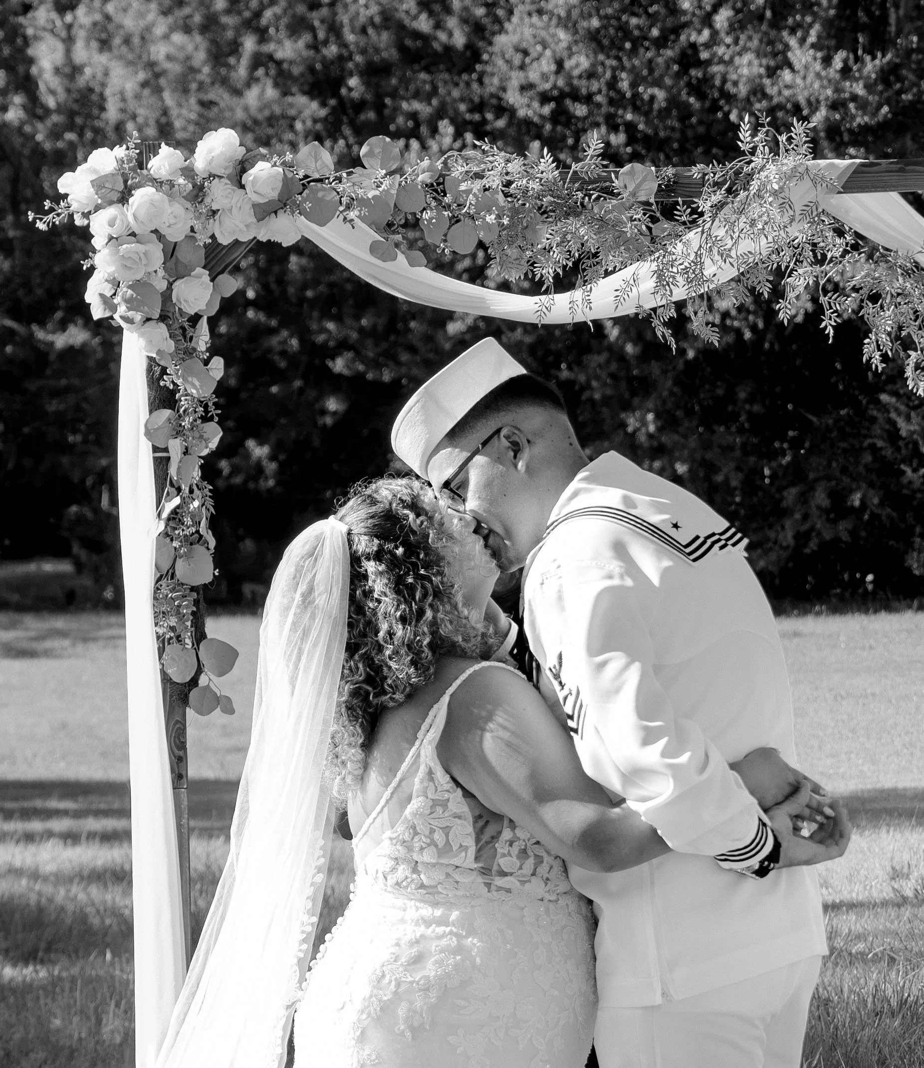 A black and white photo of a bride and groom kissing outdoors during their wedding ceremony, with a decorated archway adorned with flowers and greenery in the background.