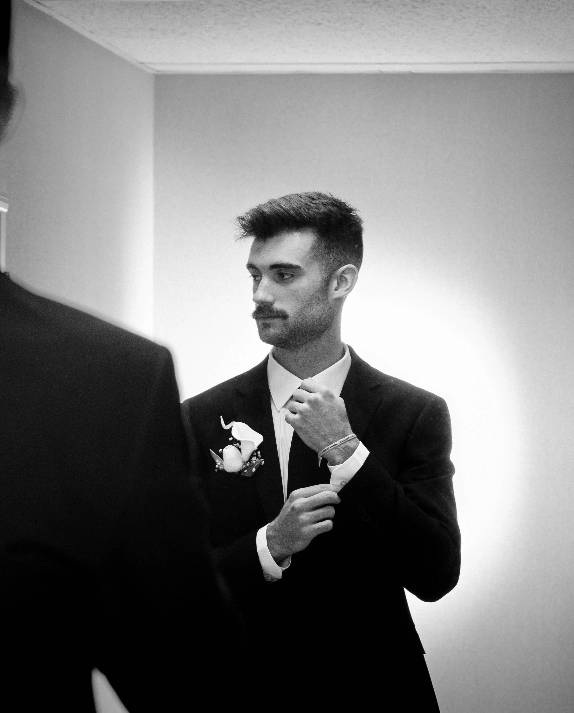 A man in a tuxedo with a boutonnière and boutonnière ribbon is adjusting his jacket while looking down, in a black and white photo.