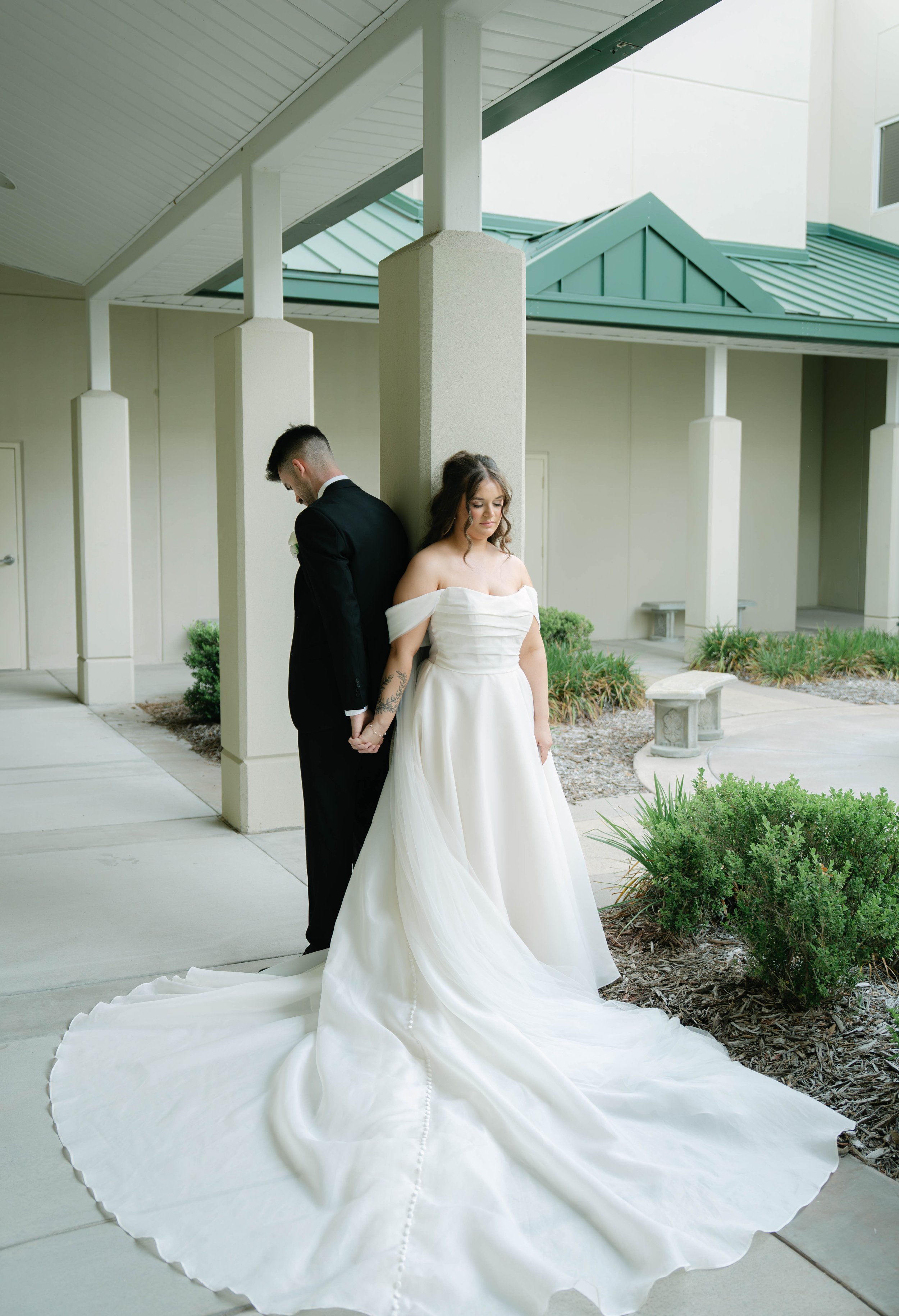 A bride and groom stand back to back outdoors under a covered walkway. The bride wears a white off-shoulder wedding gown with a long train, and the groom is dressed in a black tuxedo. They hold hands behind their backs and appear contemplative.