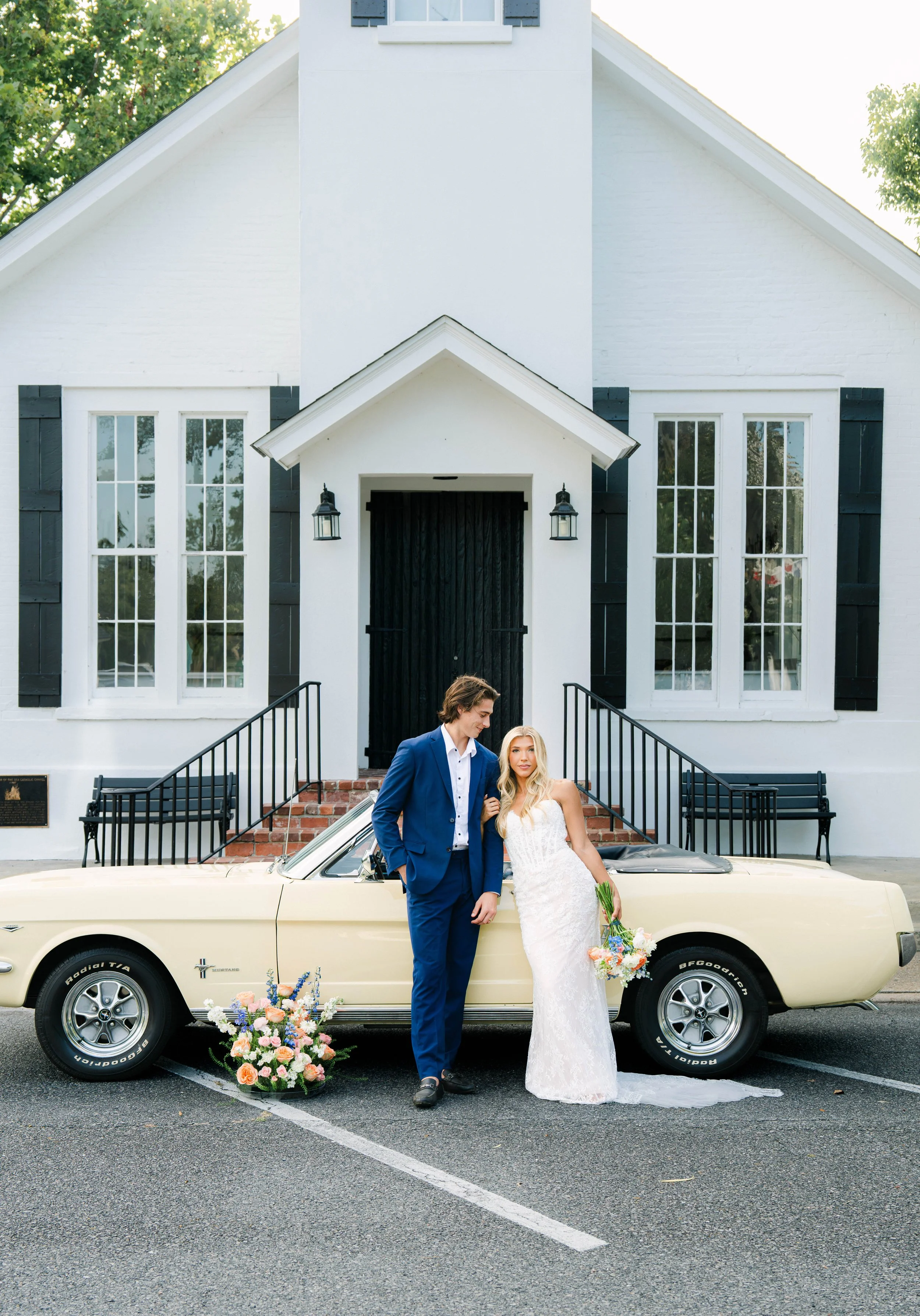 A newlywed couple standing in front of a vintage cream-colored car with pink and white flowers on the ground, in front of a white house with black accents and large windows. The groom is wearing a blue suit and the bride is wearing a white wedding dr