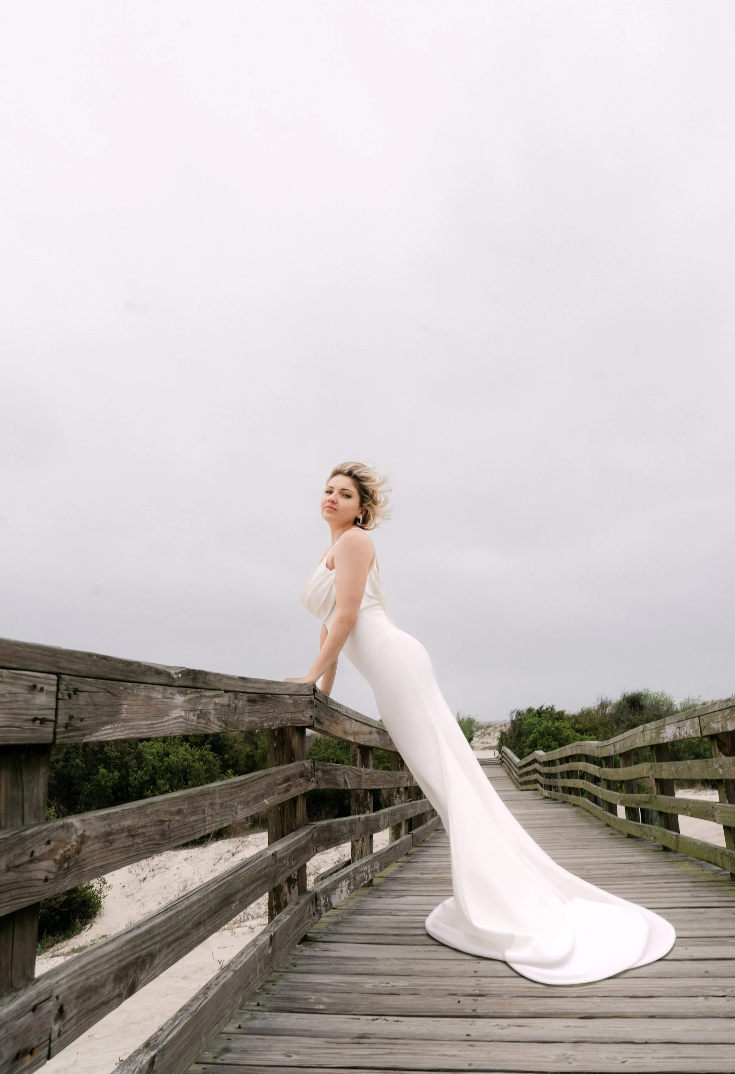 A woman in a long white dress standing on a wooden boardwalk, with her hand resting on the railing and looking at the camera.