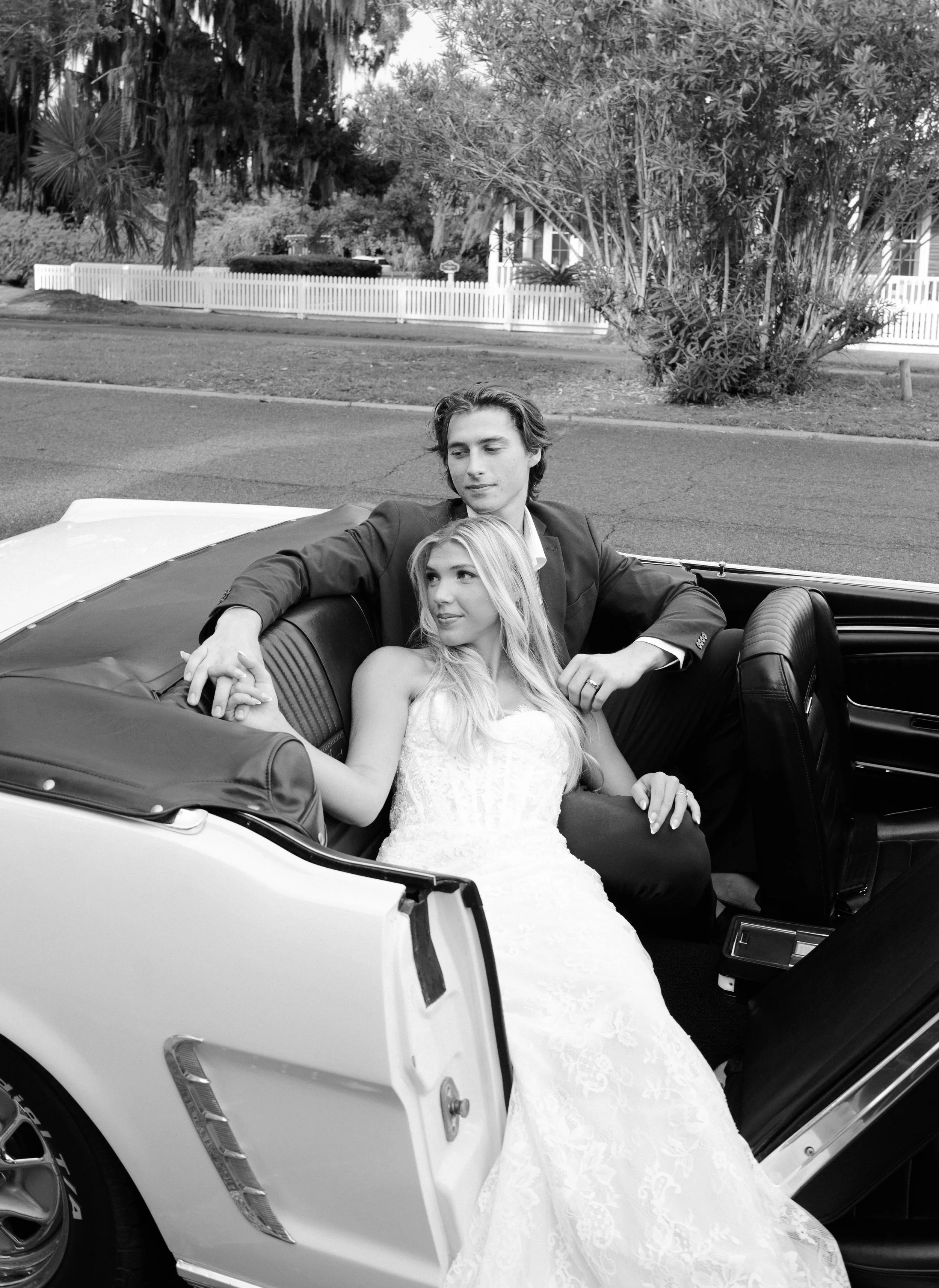 A young couple in formal wedding attire sitting in a vintage convertible car, with the bride in a white gown and the groom in a suit, on a quiet residential street with trees and a white picket fence in the background.