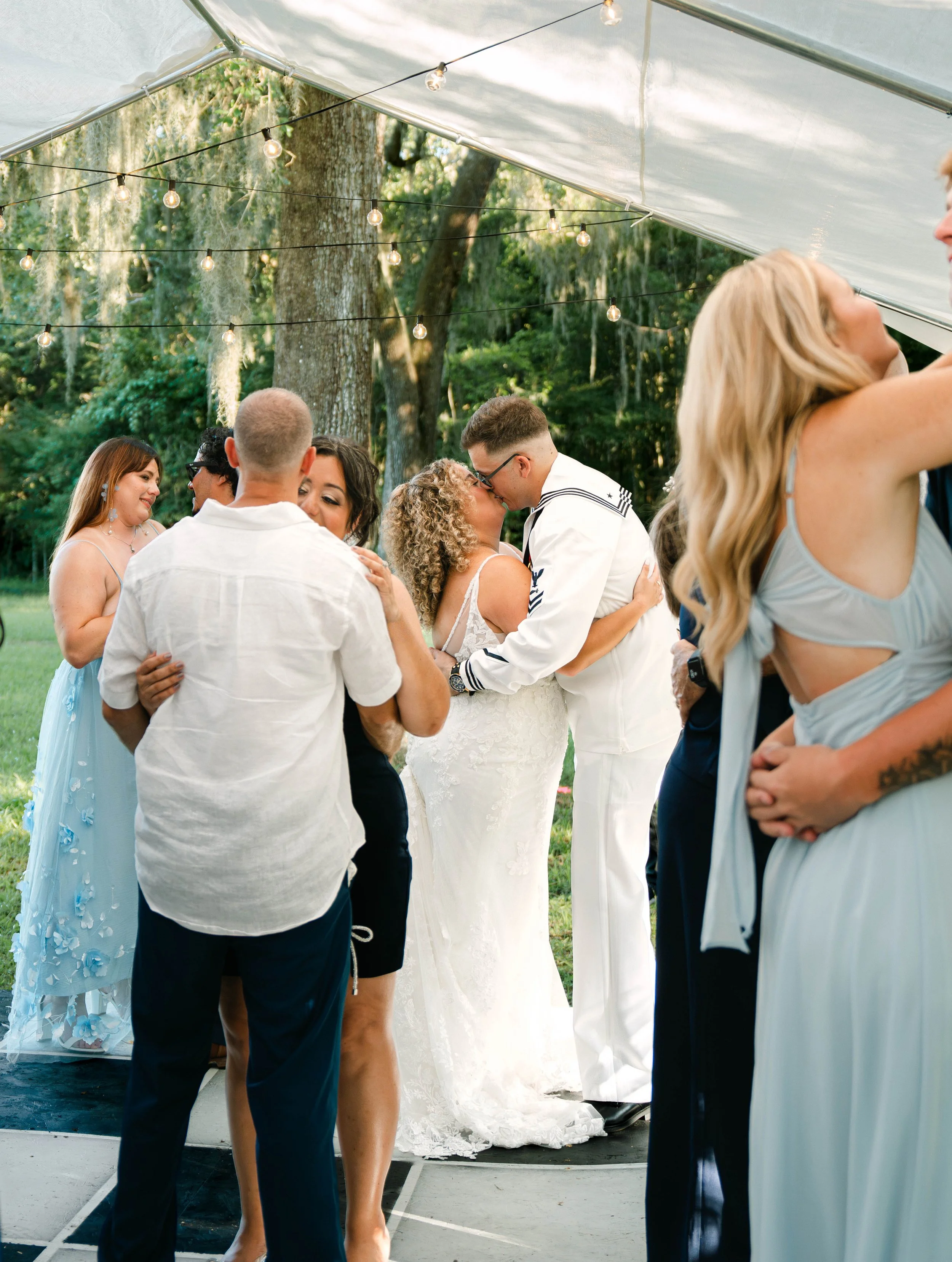 People dancing and kissing under a canopy with string lights at an outdoor event in a wooded area.