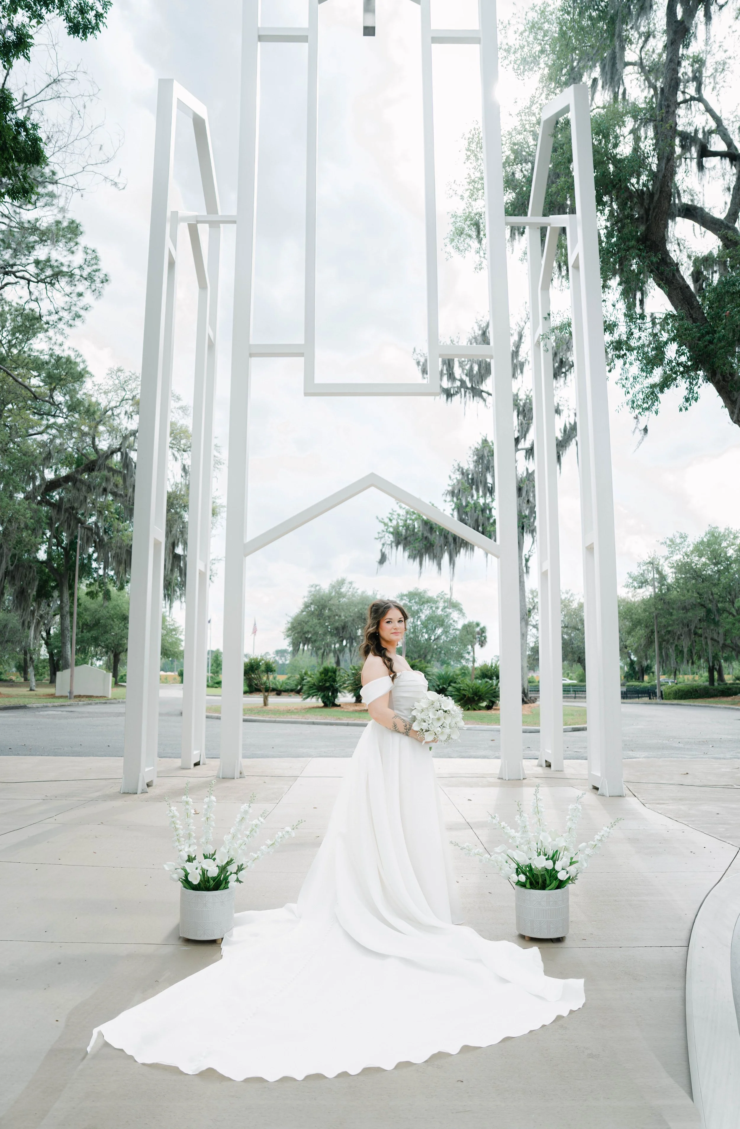 A bride in a white wedding dress holding a bouquet of white flowers standing under a modern white geometric outdoor wedding arch with white floral arrangements at its base, on a concrete platform, with green trees and cloudy sky in the background.