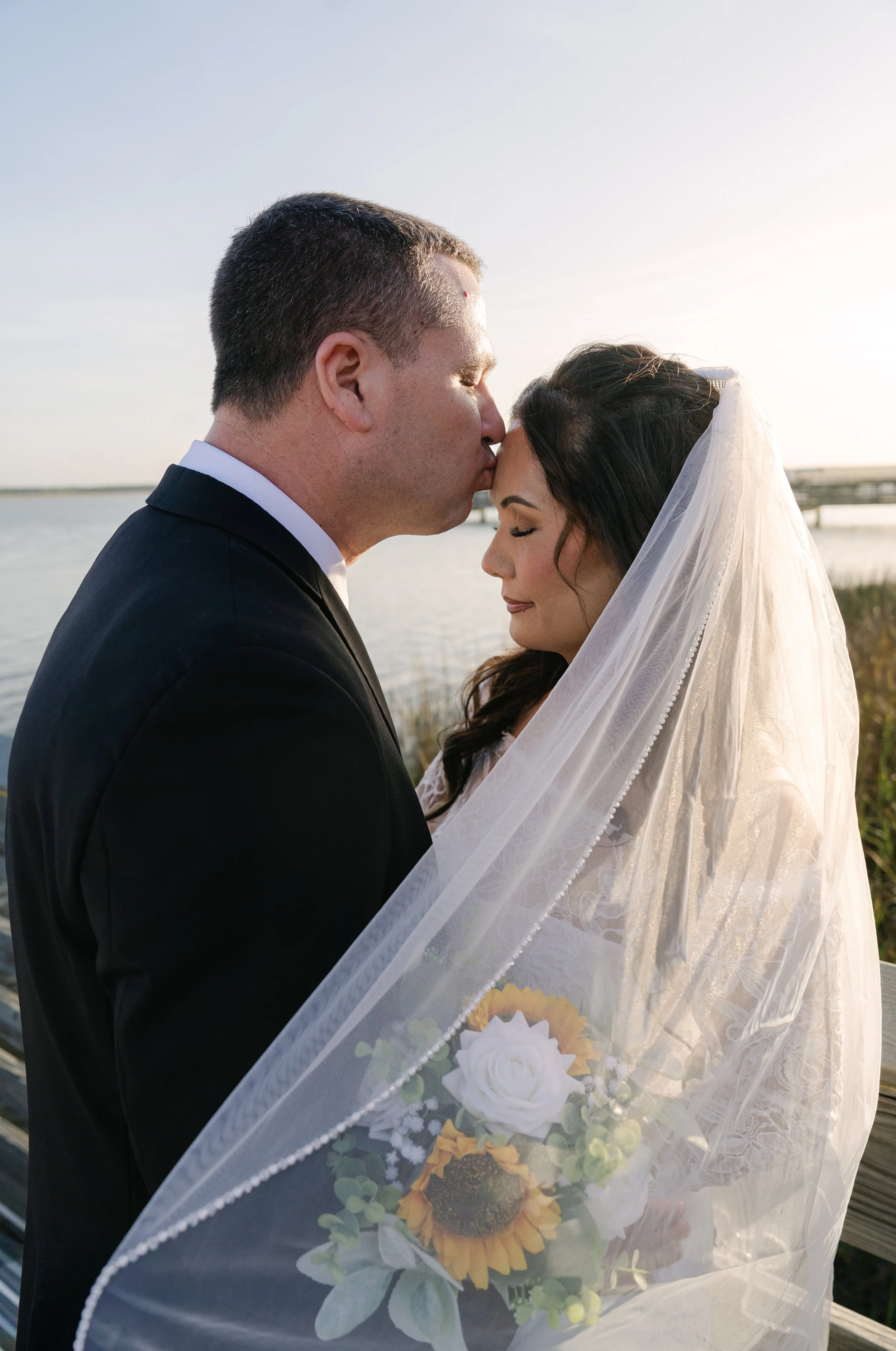 A bride and groom share a kiss outdoors near a body of water at sunset. The groom is wearing a tuxedo, and the bride is wearing a veil and lace dress holding a sunflower bouquet.