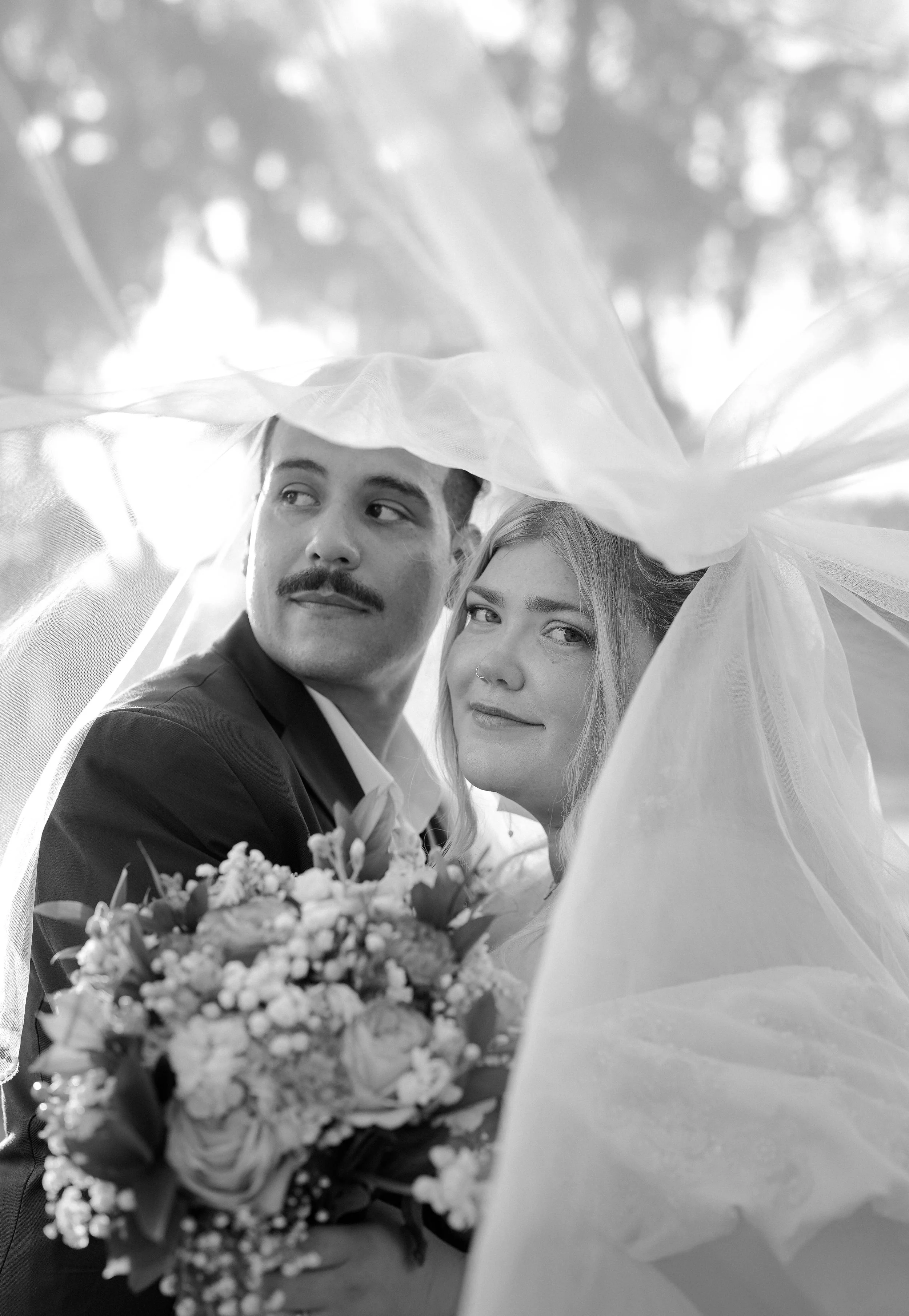 Black and white photo of a bride and groom under a veil with a bride holding a bouquet of flowers.