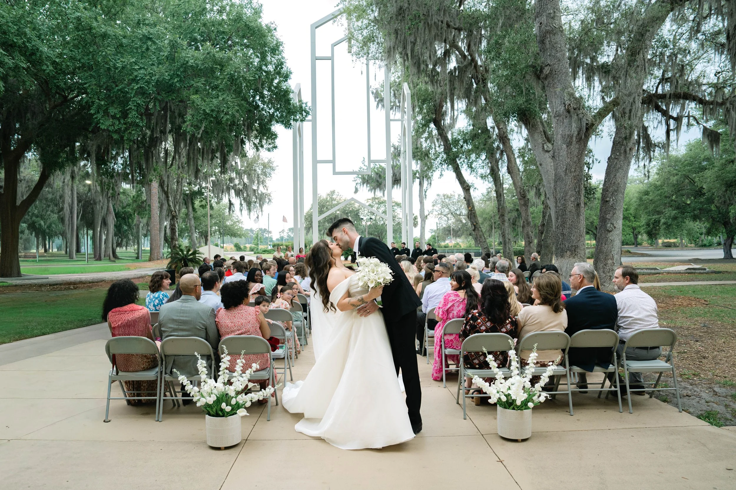 A bride and groom sharing a kiss during their outdoor wedding ceremony, with guests seated on chairs and trees in the background.
