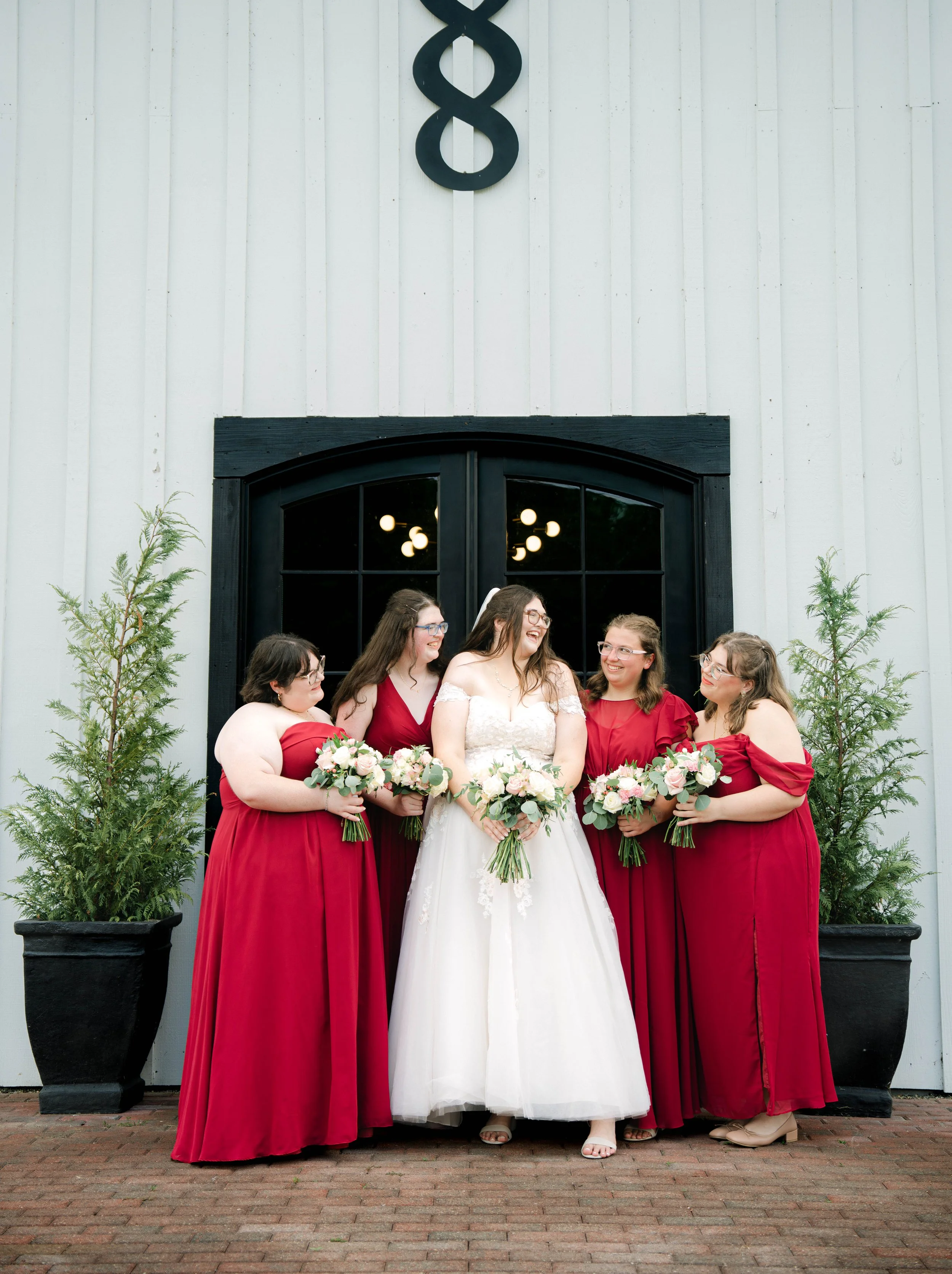 A bride in a white wedding dress surrounded by six bridesmaids in red dresses, all holding bouquets, standing in front of a white wall with black window and an ampersand sign above.