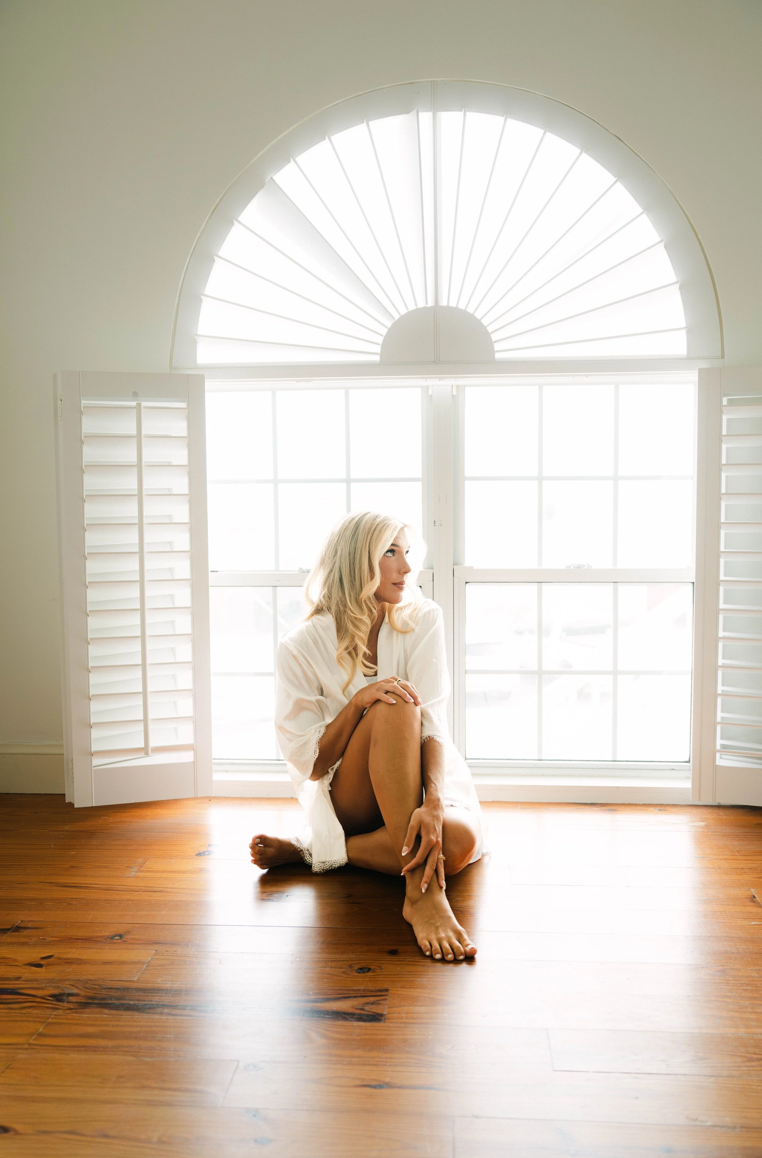 A woman with blonde hair sitting on a wooden floor in front of a large window with white shutters, wearing a white robe and black stockings, looking to the side.