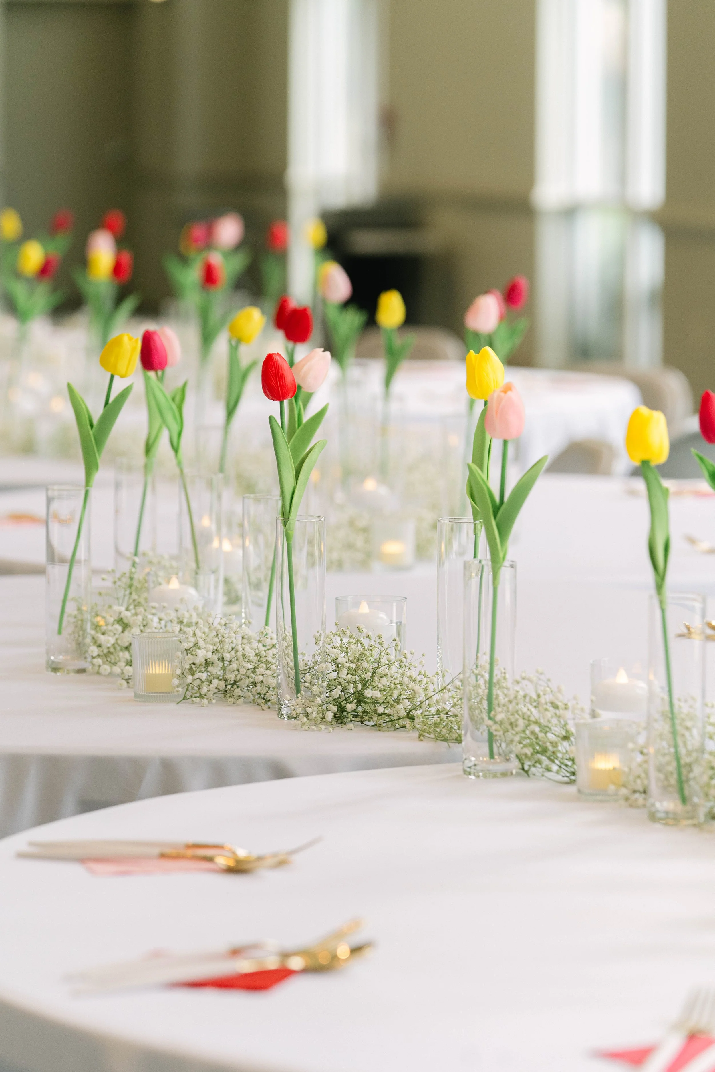Table decorated with colorful tulips in glass vases, surrounded by small candles and white floral accents in a bright room.