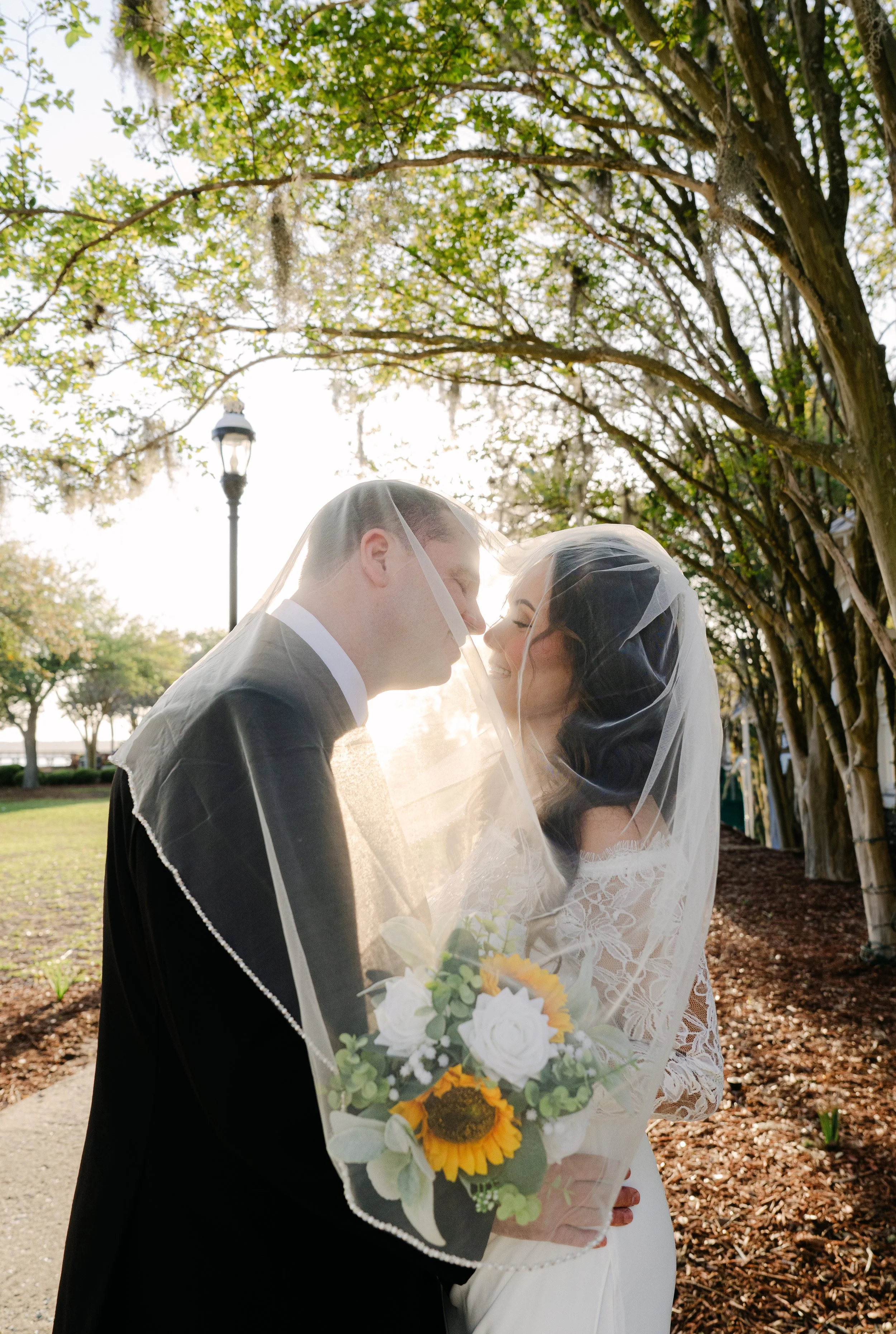 A bride and groom nose to nose under a veil outdoors on a sunny day, with trees and a lamp post in the background.