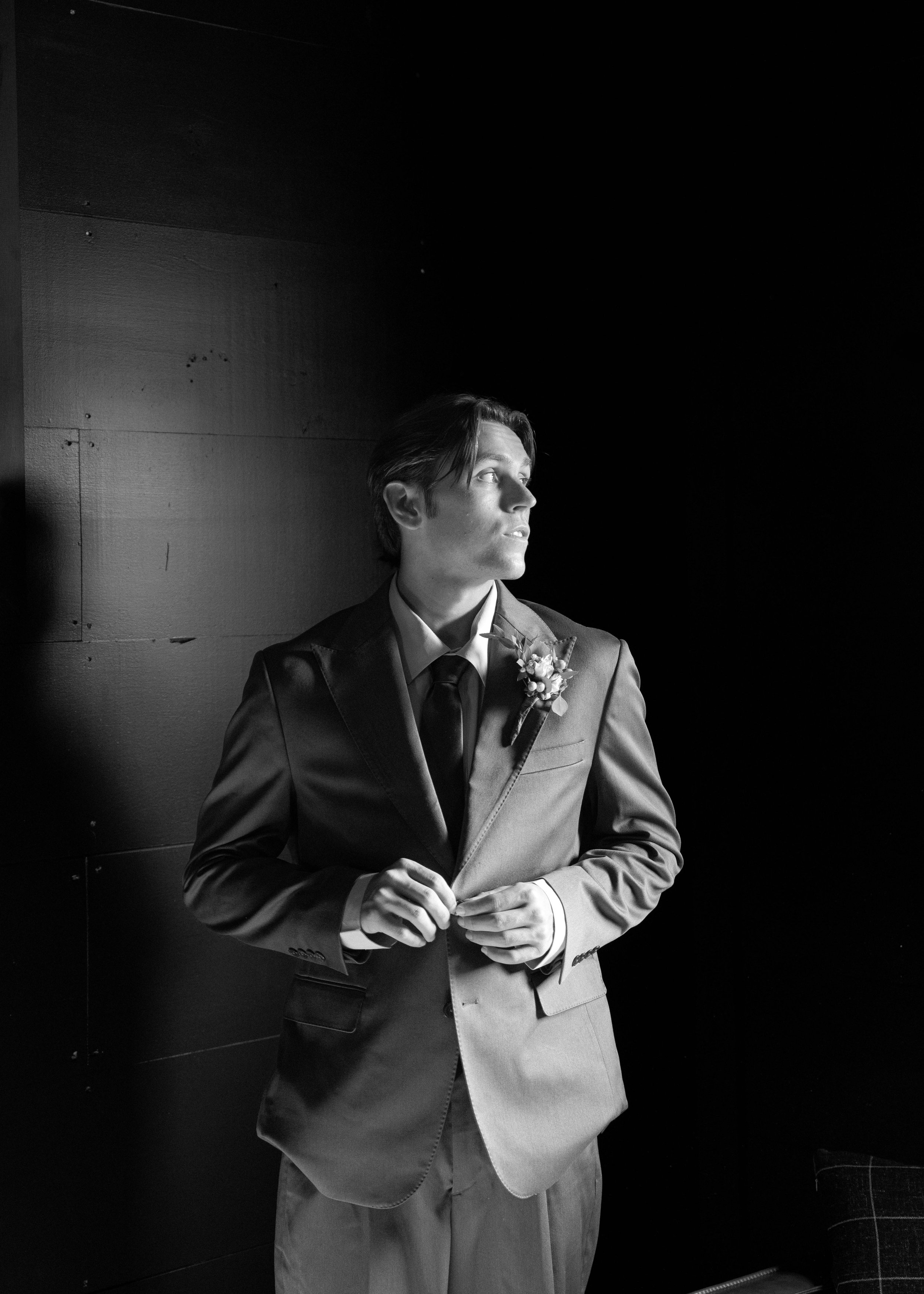 Black and white photo of a man in a suit with a flower boutonniere, looking to the side, standing against a dark wall with some visible wood paneling.