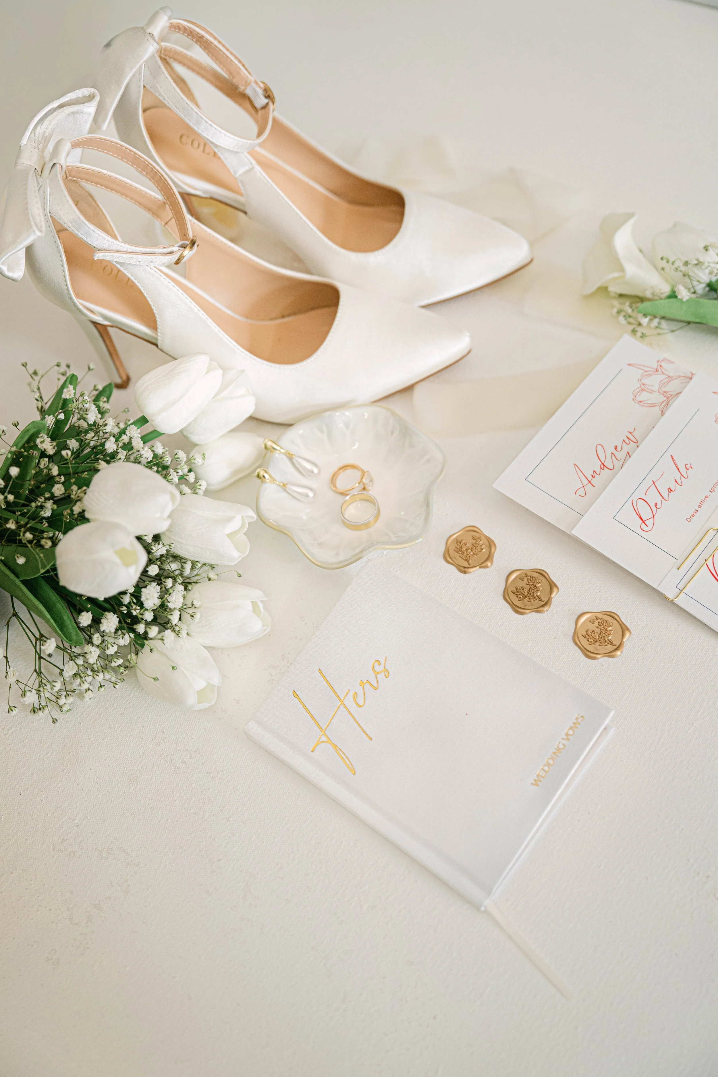 A pair of white high heel shoes with ankle straps, a bouquet of white tulips and baby's breath flowers, wedding invitations with cursive writing, gold rings, and wax seals on a white surface.