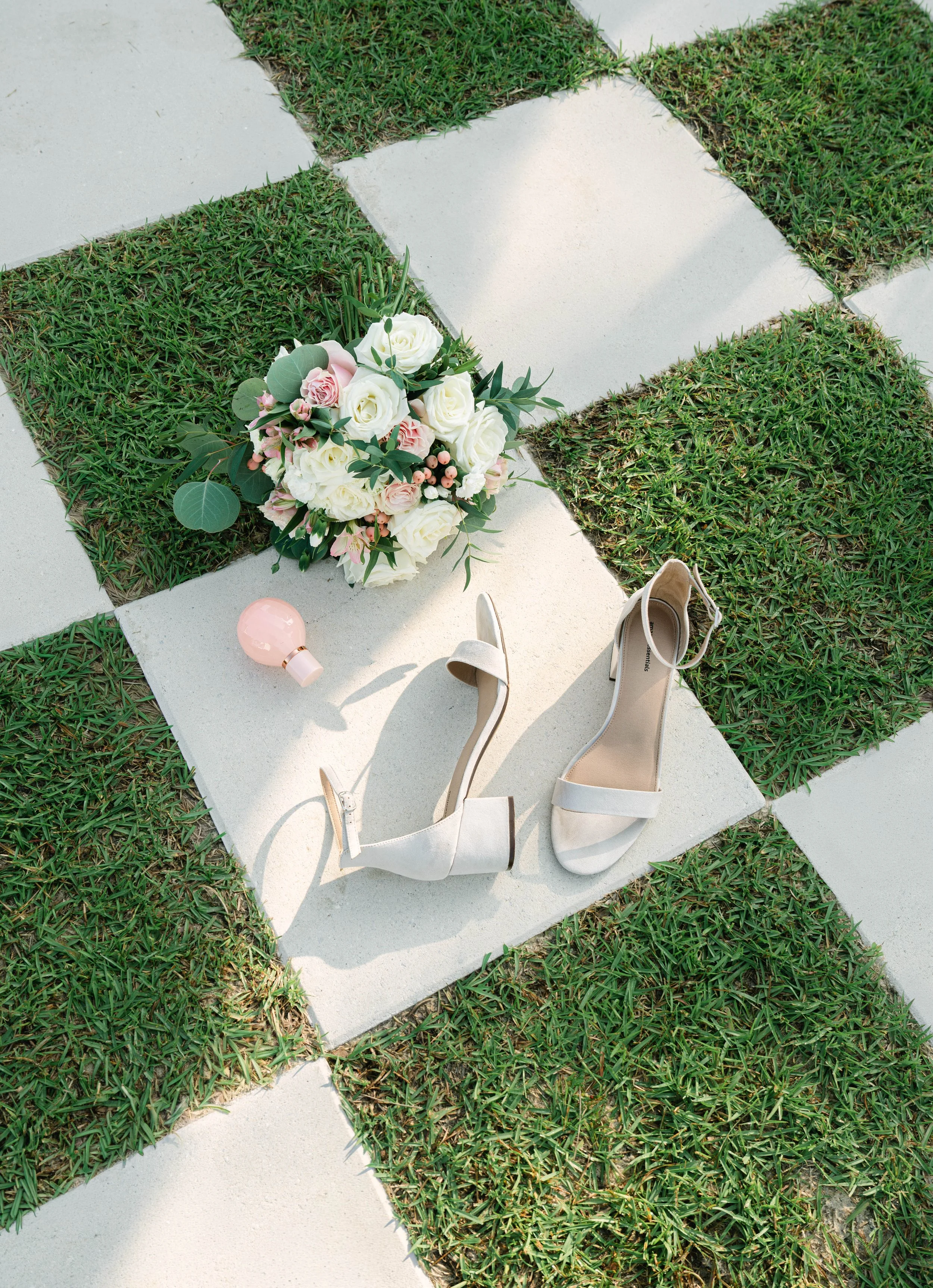 A bouquet of white and pink roses with greenery, a pair of beige high-heeled shoes, and a pink lamp placed on a white tile pathway surrounded by grass.