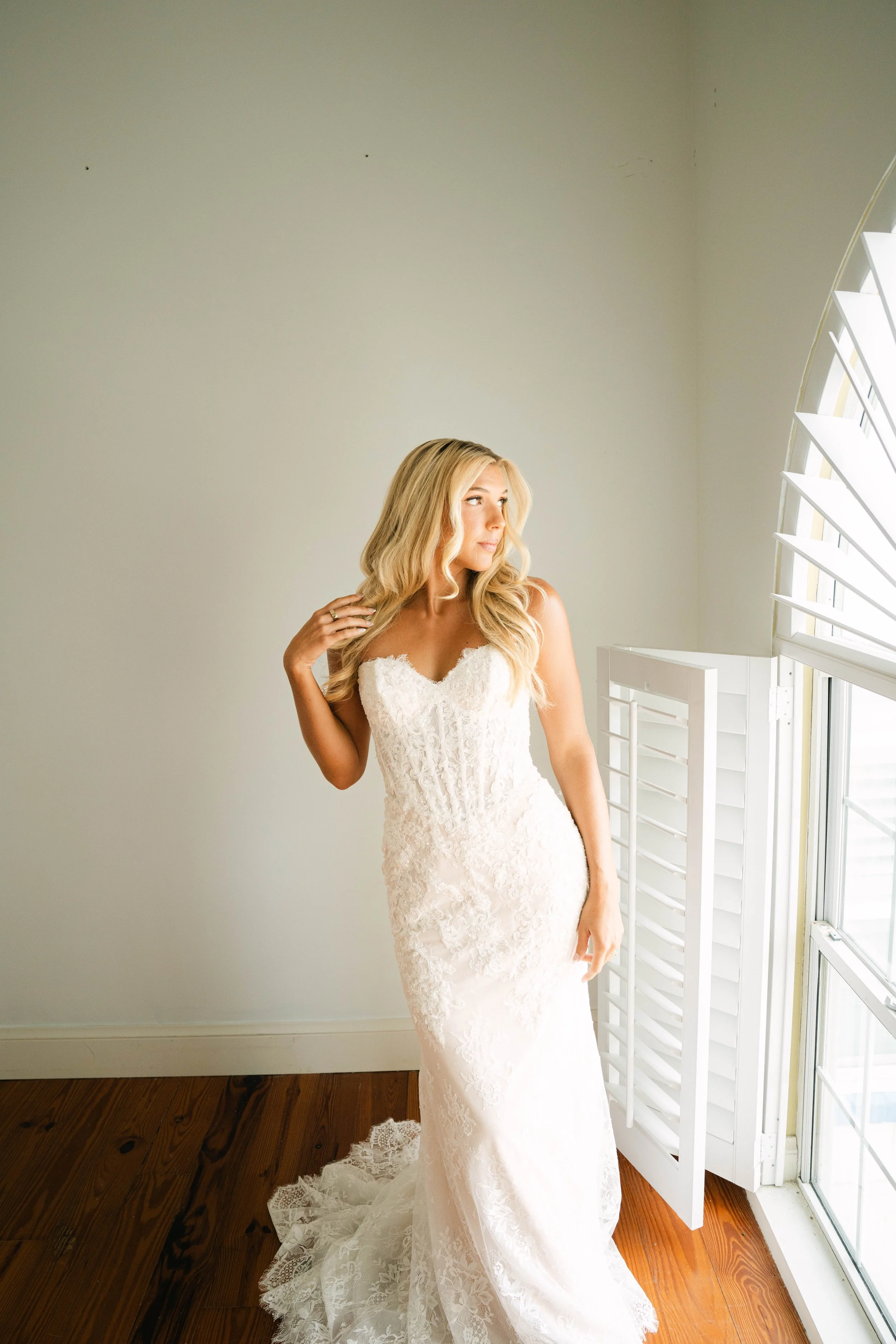 A woman in a white lace wedding dress standing near a window with open white shutters, looking thoughtfully to the side, with blonde hair and a gentle expression.