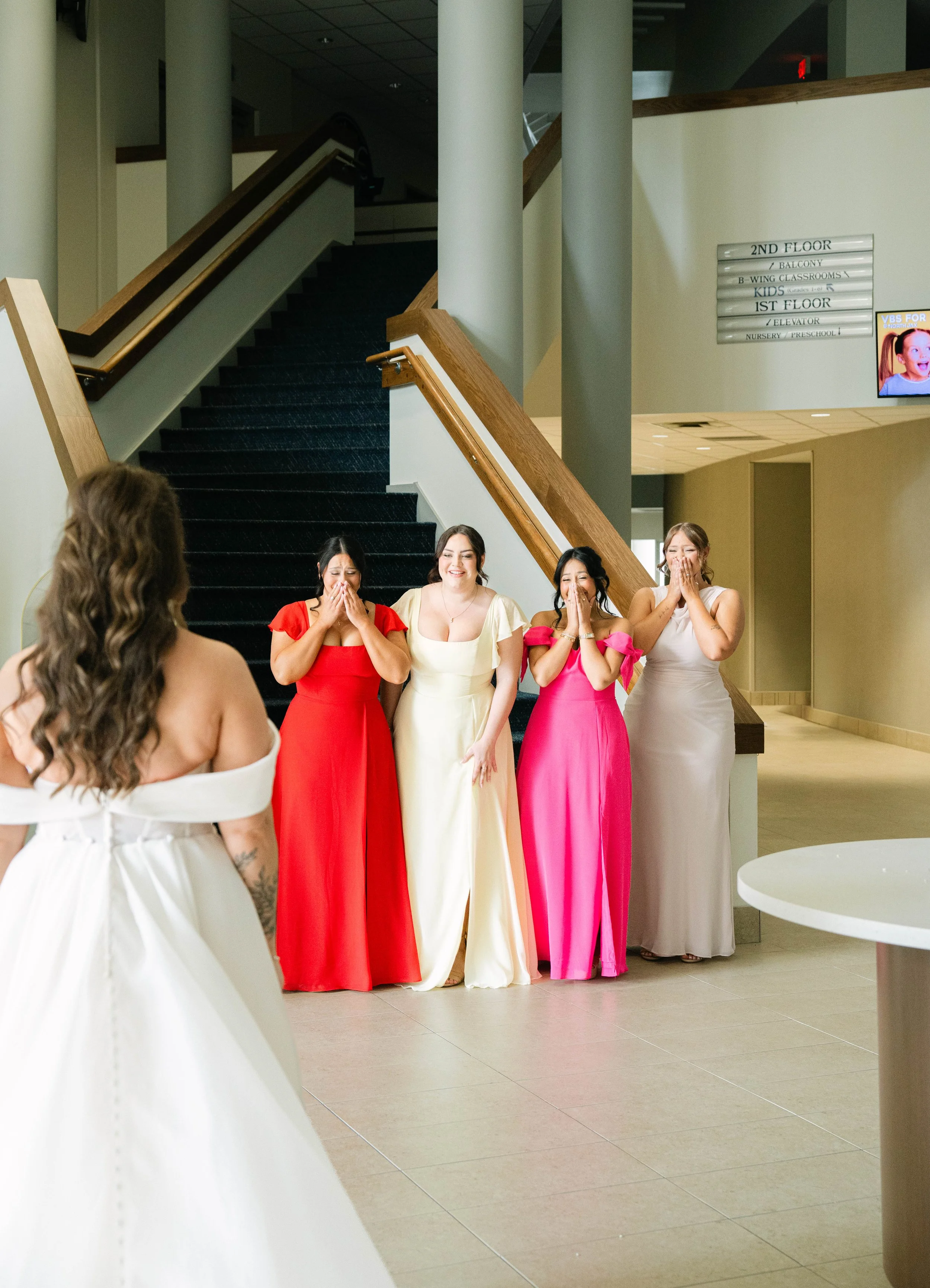 A bride in a white off-shoulder dress is facing four women in vibrant dresses, who are reacting happily with surprise and joy, inside a building with a staircase and directory sign on the wall.