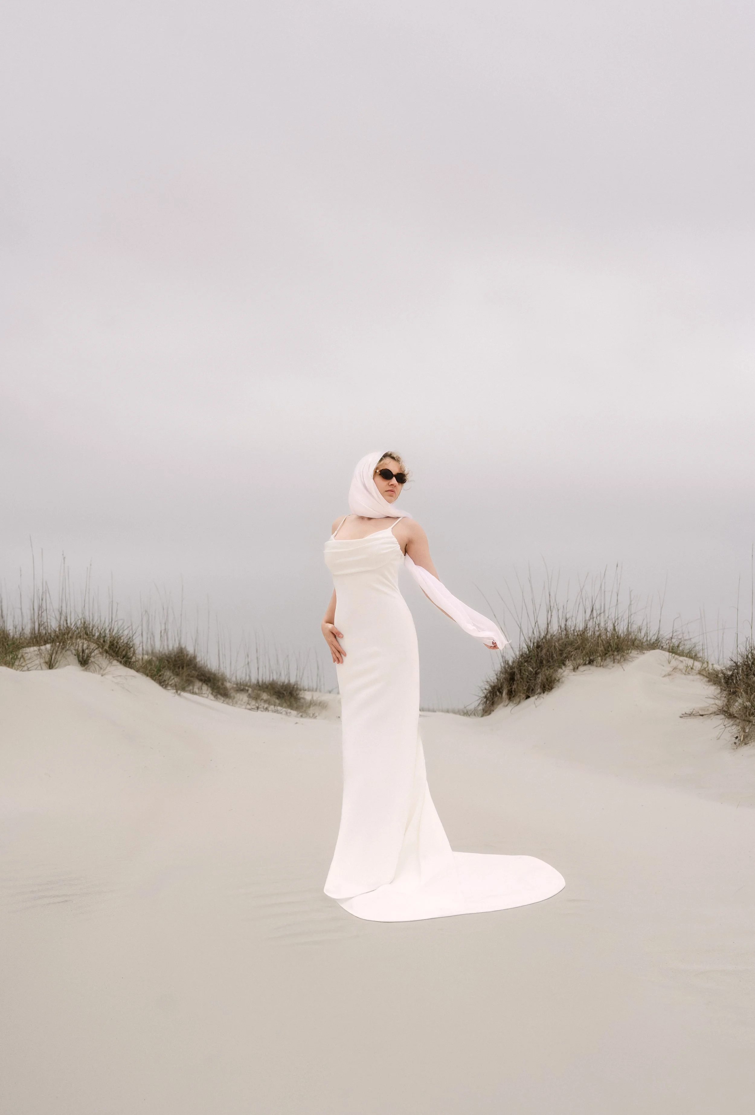 A woman in a long white dress and black sunglasses standing on a sandy beach with dry grasses and a foggy, gray sky in the background.