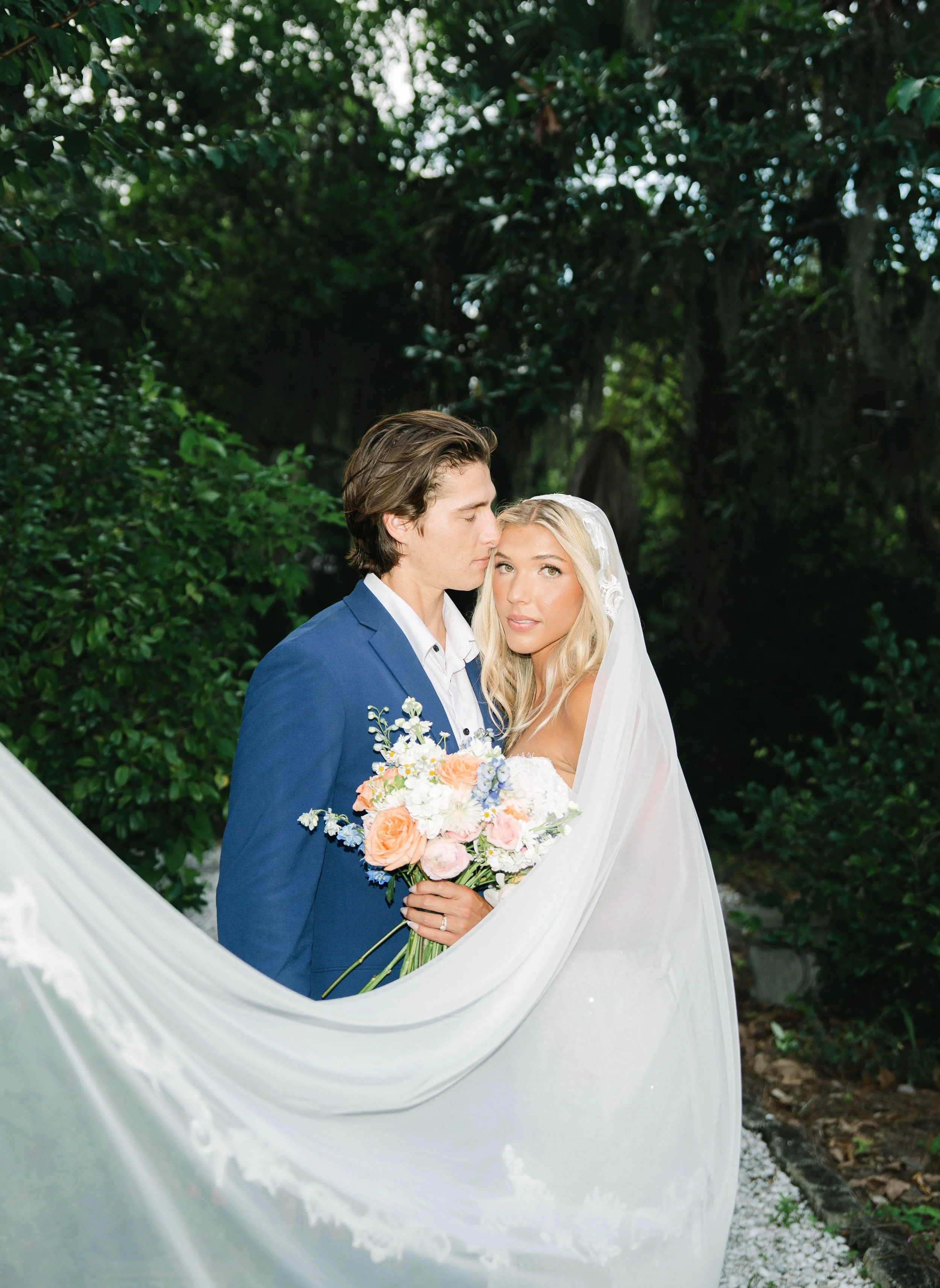 A bride and groom standing together outdoors, with the groom kissing the bride on the forehead, surrounded by lush greenery. The bride holds a bouquet of pastel-colored flowers and wears a white veil and dress, while the groom wears a blue suit and w
