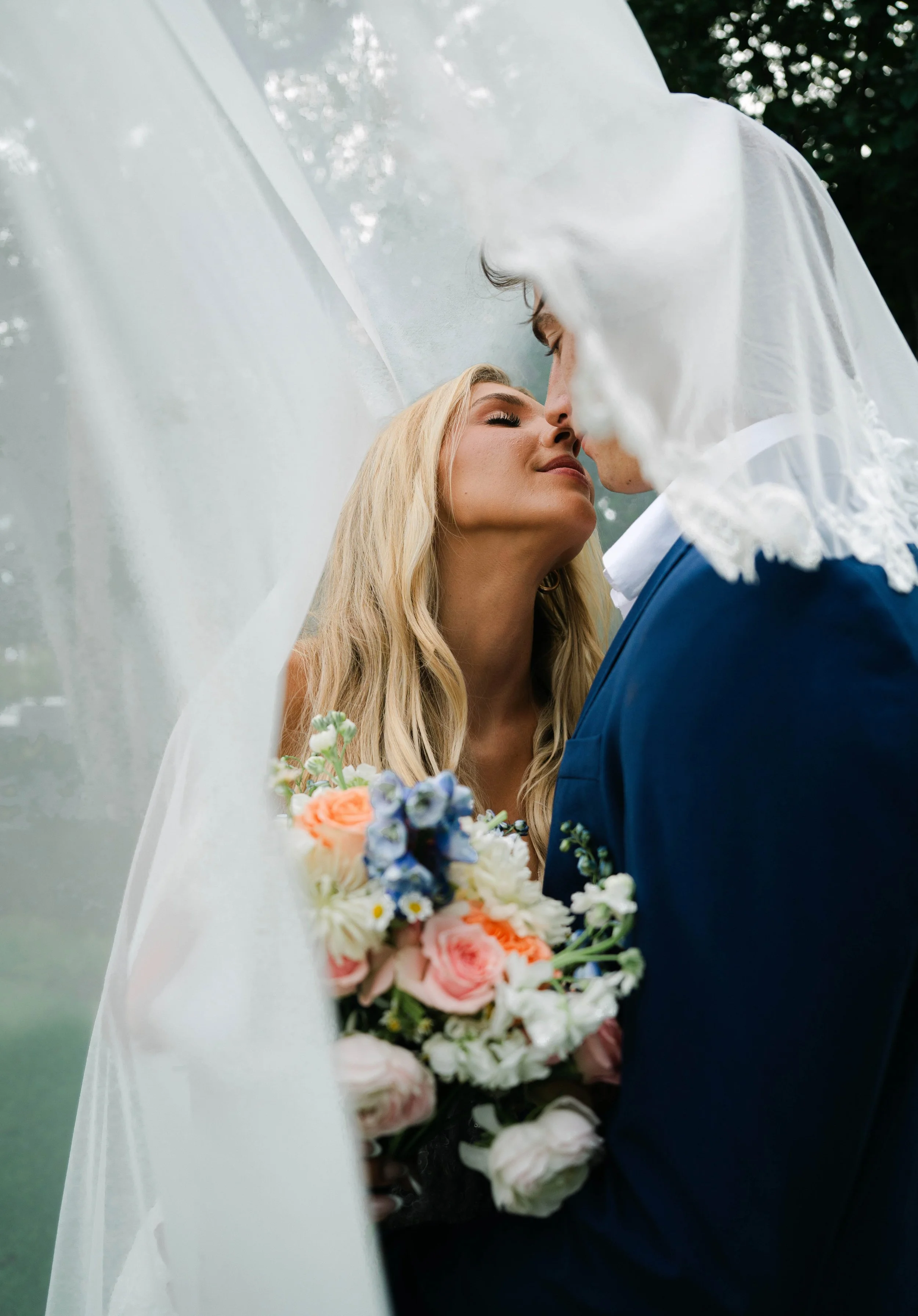 A bride and groom sharing a kiss behind a wedding veil, with the bride holding a bouquet of colorful flowers.