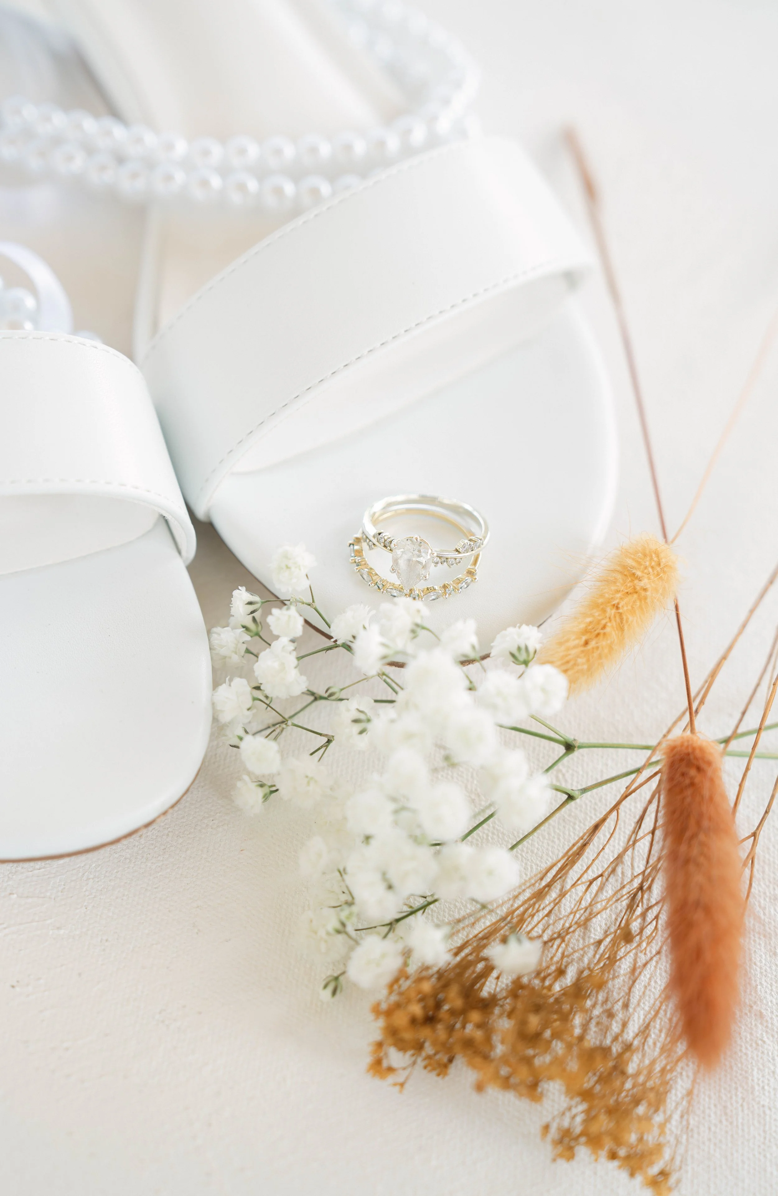 A pair of white bridal shoes with pearl details, a silver engagement ring featuring a large central stone, and small white flowers, all arranged on a light-colored surface with dried orange and beige grasses.
