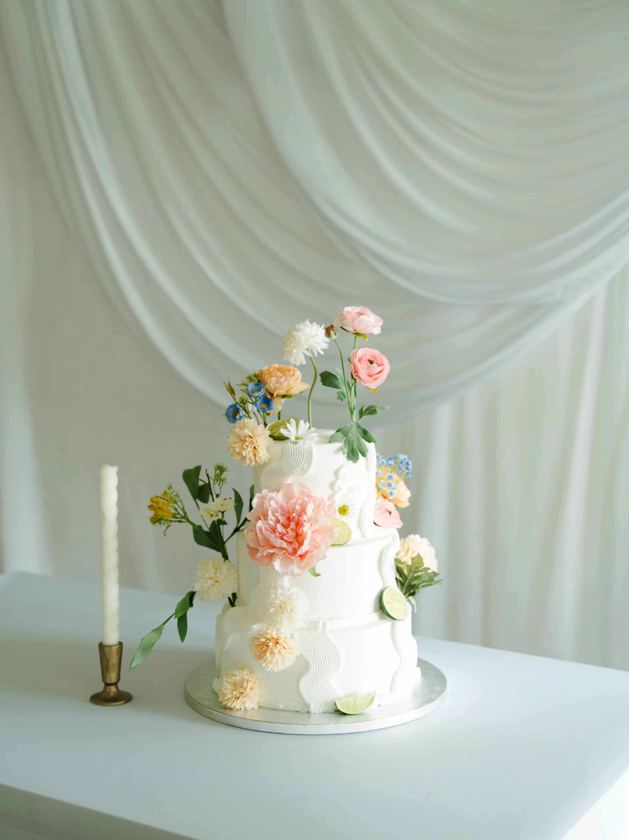 A white wedding cake decorated with various pastel-colored flowers and lime slices, topped with a small bouquet of flowers, with two tall white candles on gold bases beside it, against a backdrop of draped white fabric.