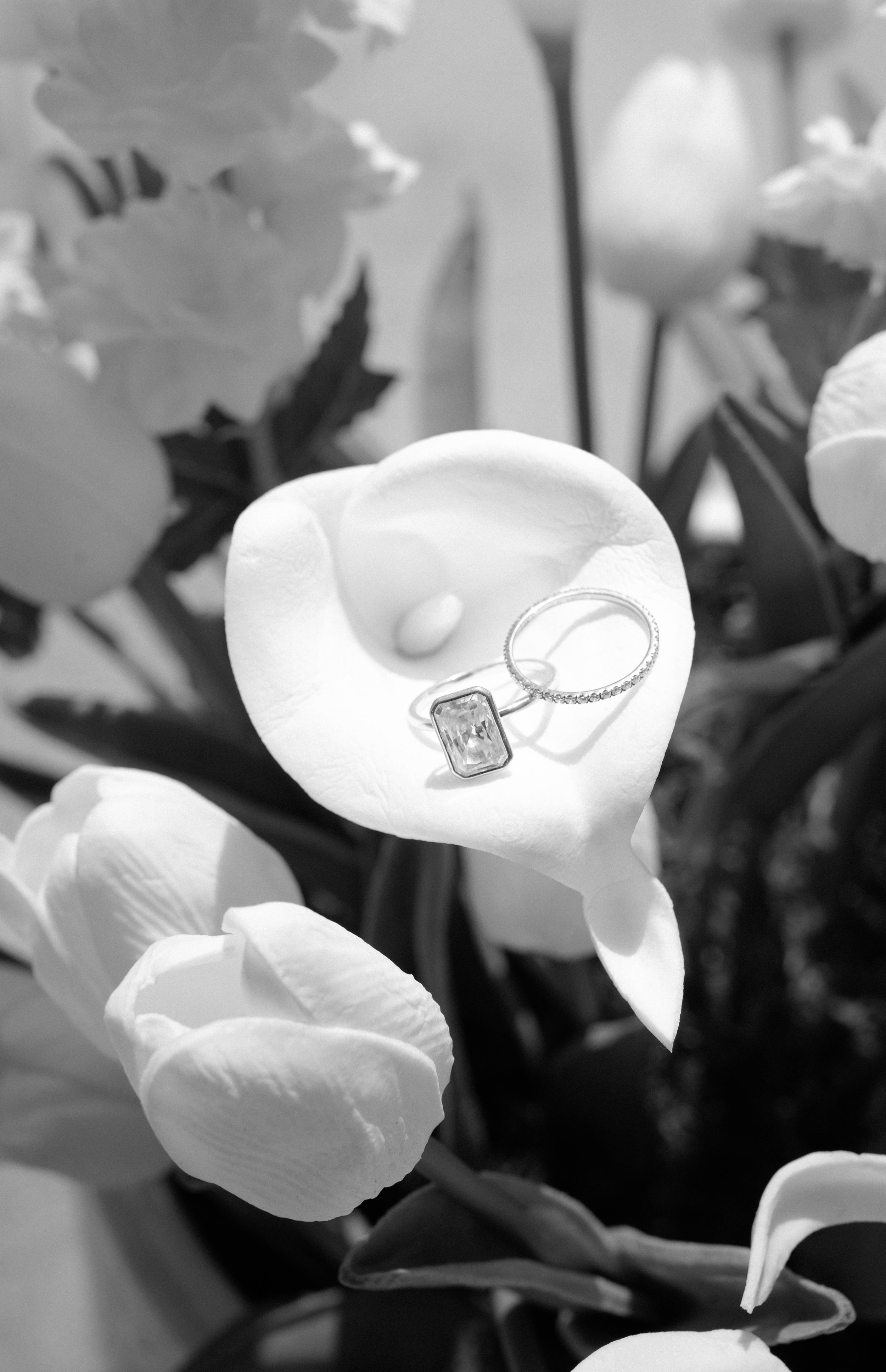 Close-up of a white calla lily flower with a diamond ring and a gemstone ring resting on its spathe, surrounded by blurred flowers in the background.