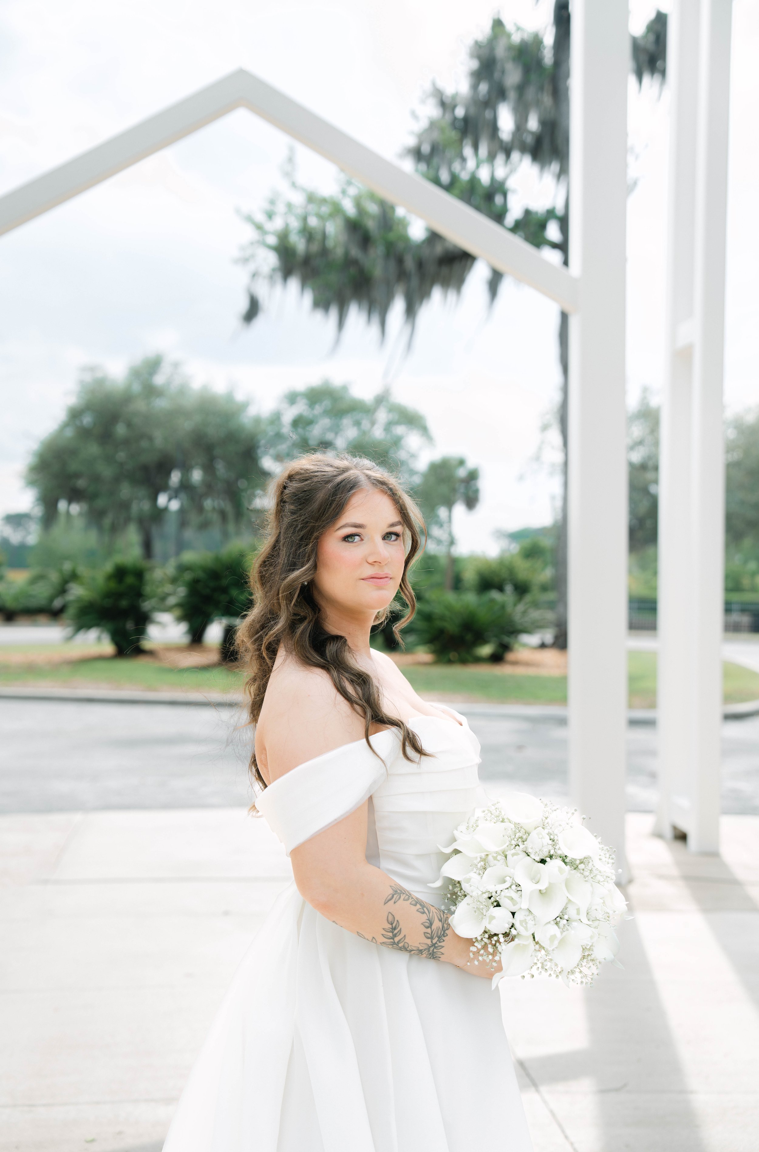 A bride with long, wavy brown hair and a floral tattoo on her left arm, dressed in an off-the-shoulder white wedding gown, standing outdoors with green trees and cloudy sky in the background, holding a bouquet of white flowers.