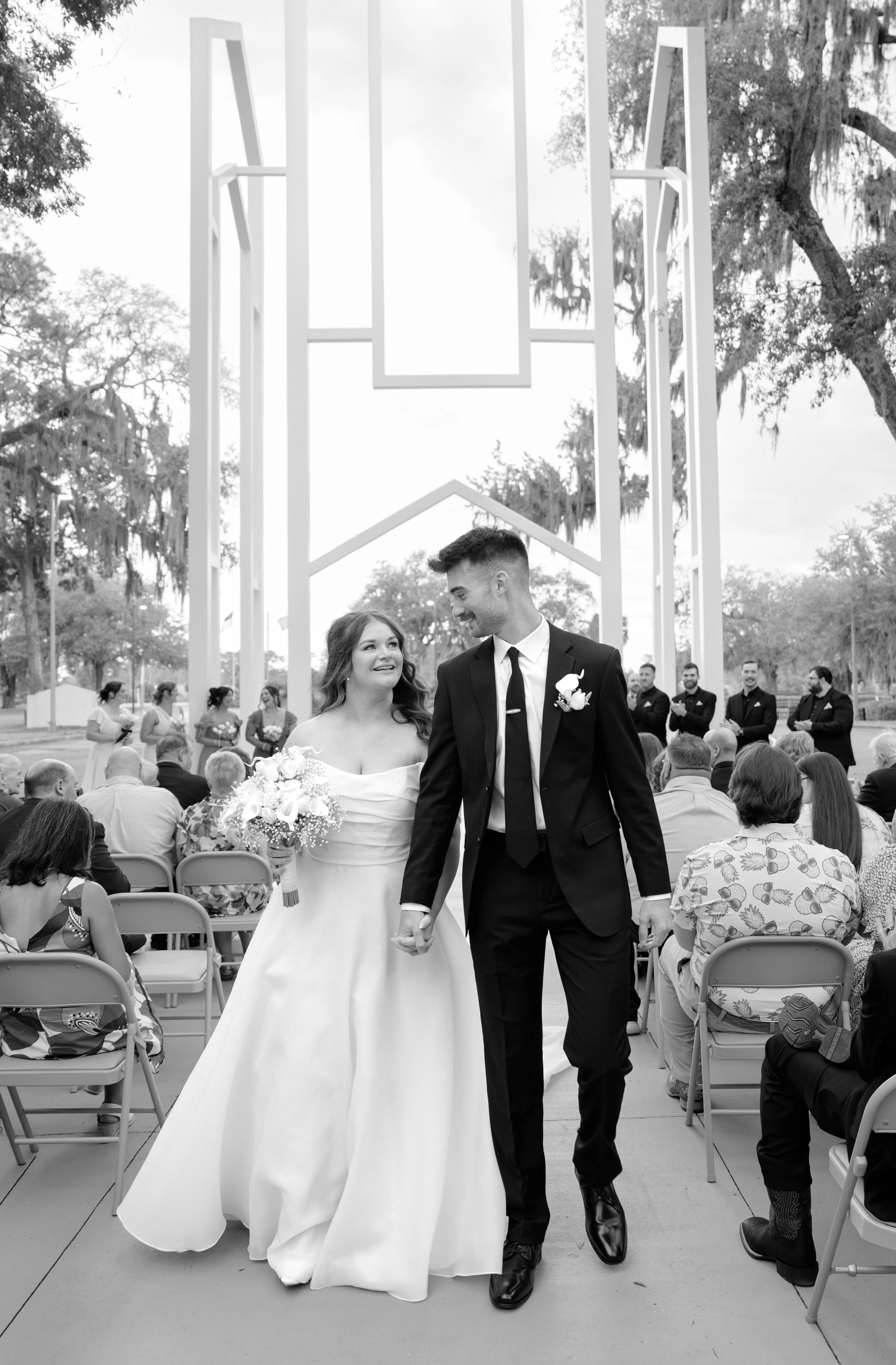 A bride and groom holding hands and smiling during their outdoor wedding ceremony, with guests seated and a large decorative frame in the background.