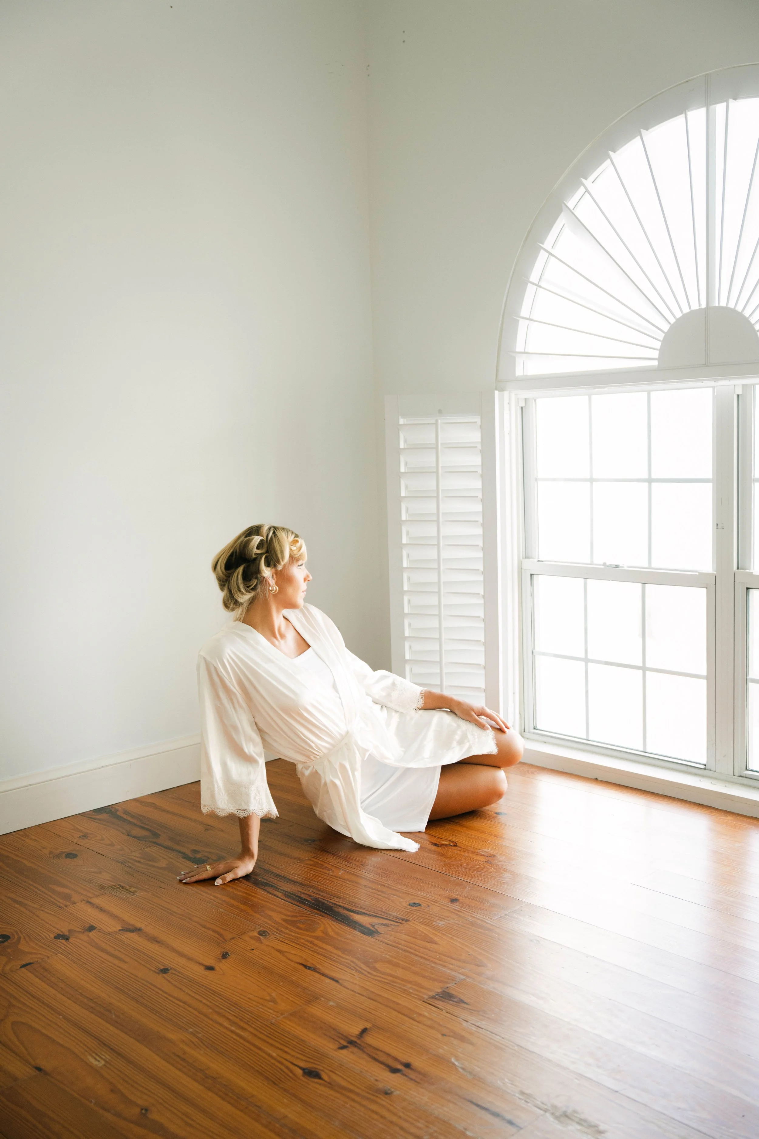 A woman with vintage styled hair posing in a white lace dress, sitting on the wooden floor near a large window with shutters, in a bright room.
