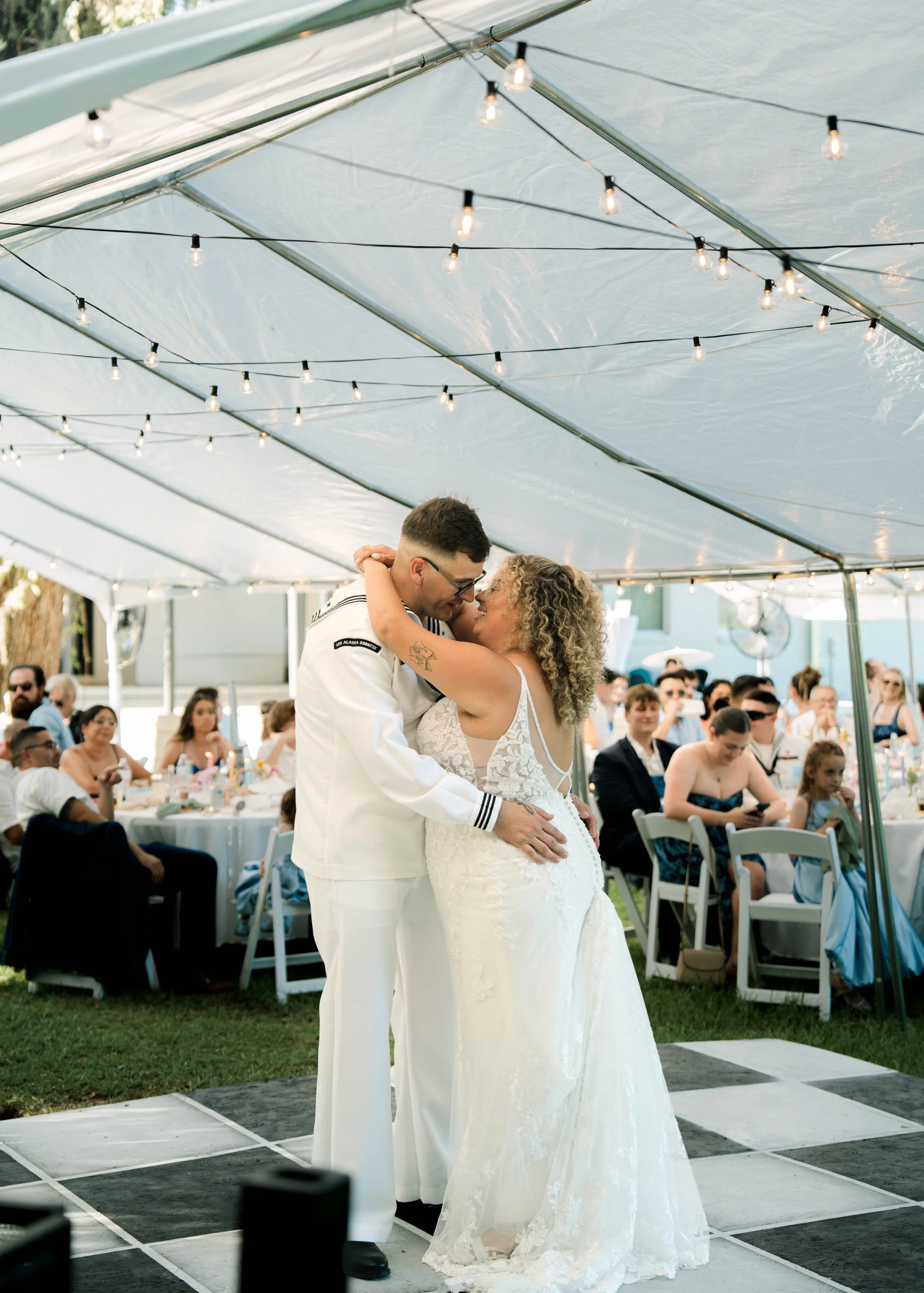 A bride and groom dance together at their wedding reception, under a canopy with string lights, in front of seated guests at outdoor tables.