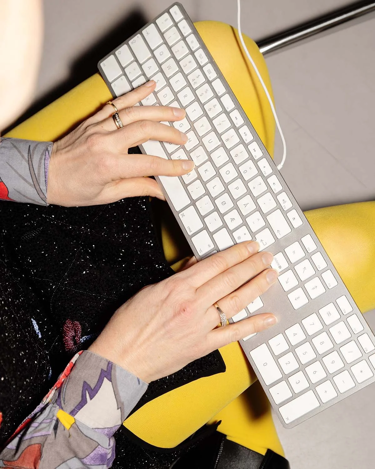 Nærbilde av en person som bruker et hvitt tastatur, som ligger på personens knær, med en gul stol og en trådløs kabel i bakgrunnen.