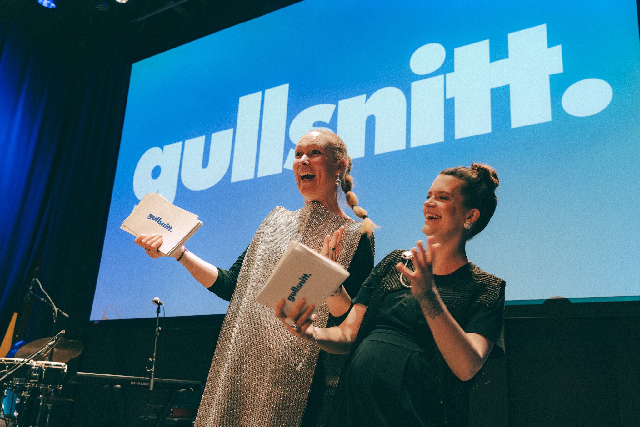 To kvinner på scenen under en forestilling, den ene holder en plastmappe med teksten 'gullsint', og bak dem vises et prosjektor med et blått lerret med stor tekst.
