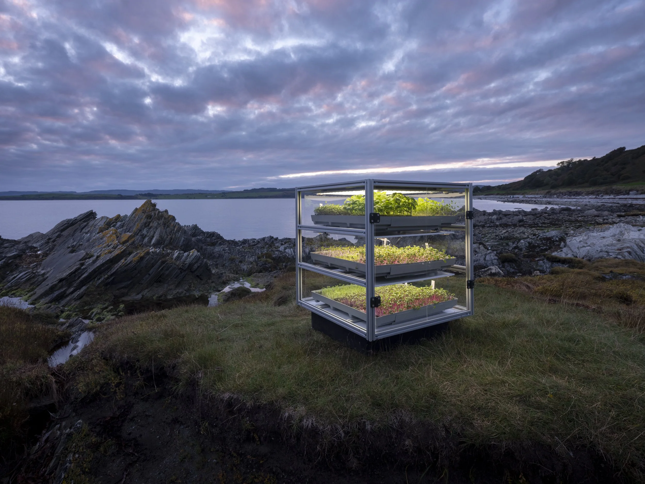 A three-tiered indoor garden in a transparent enclosure situated outdoors on rocky terrain near a body of water, with cloudy sky overhead.