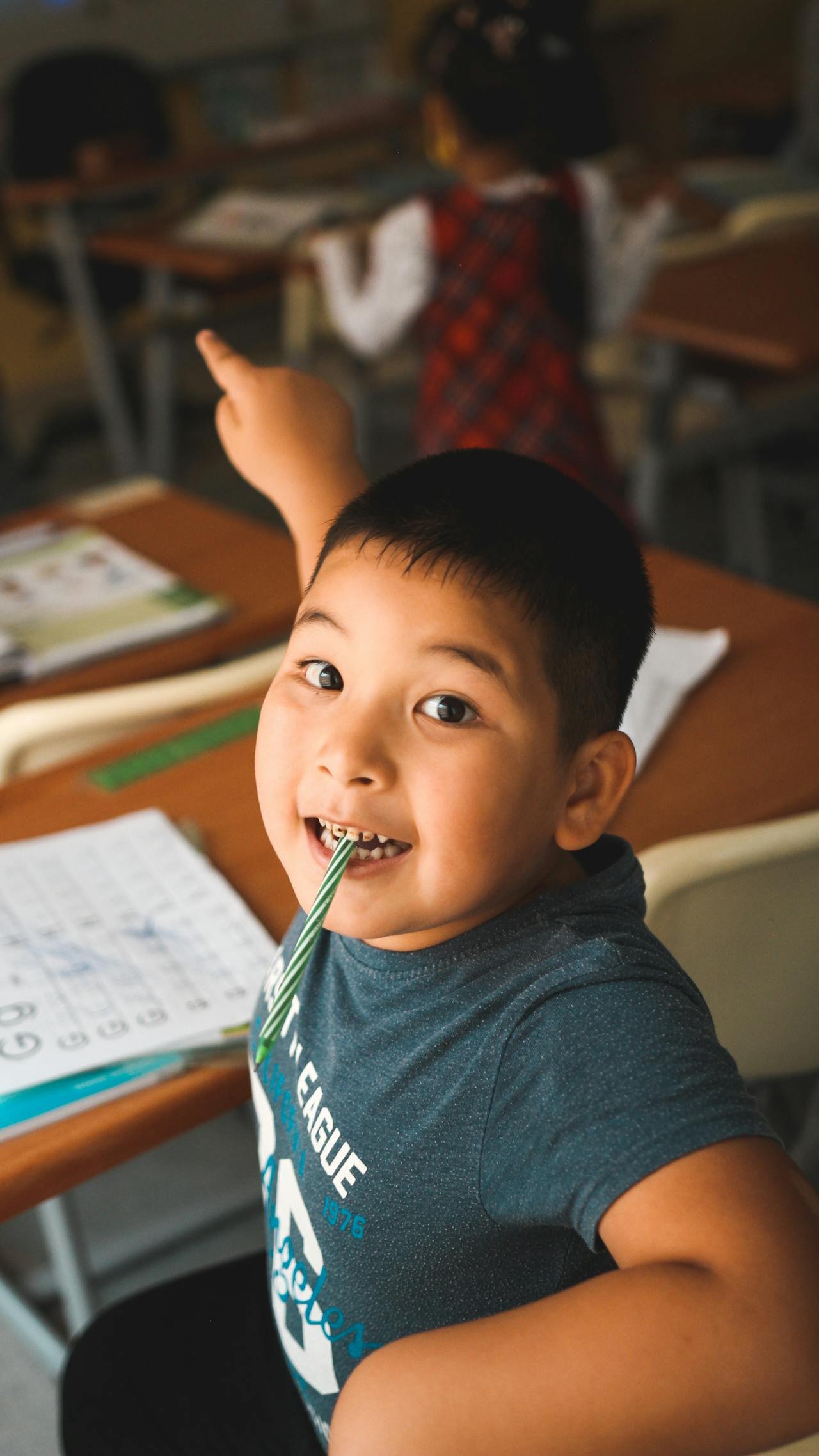Young boy in a classroom holding a green-striped straw in his mouth, smiling happily at the camera, with schoolbooks and worksheets on his desk.