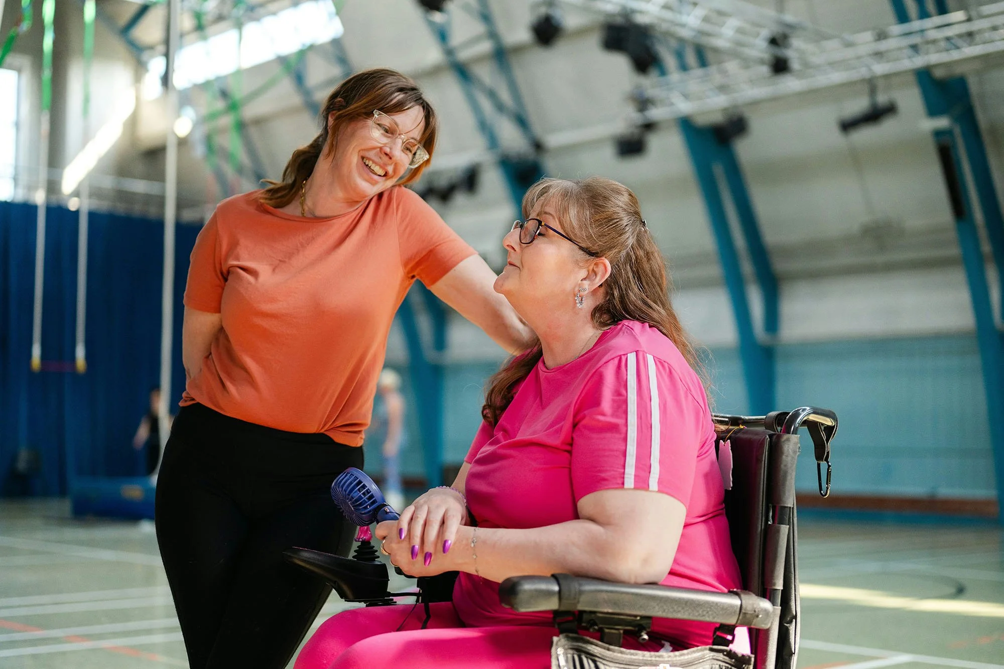 A woman in an orange shirt smiles and leans toward a woman in a pink shirt who is sitting in a wheelchair in a gymnasium.