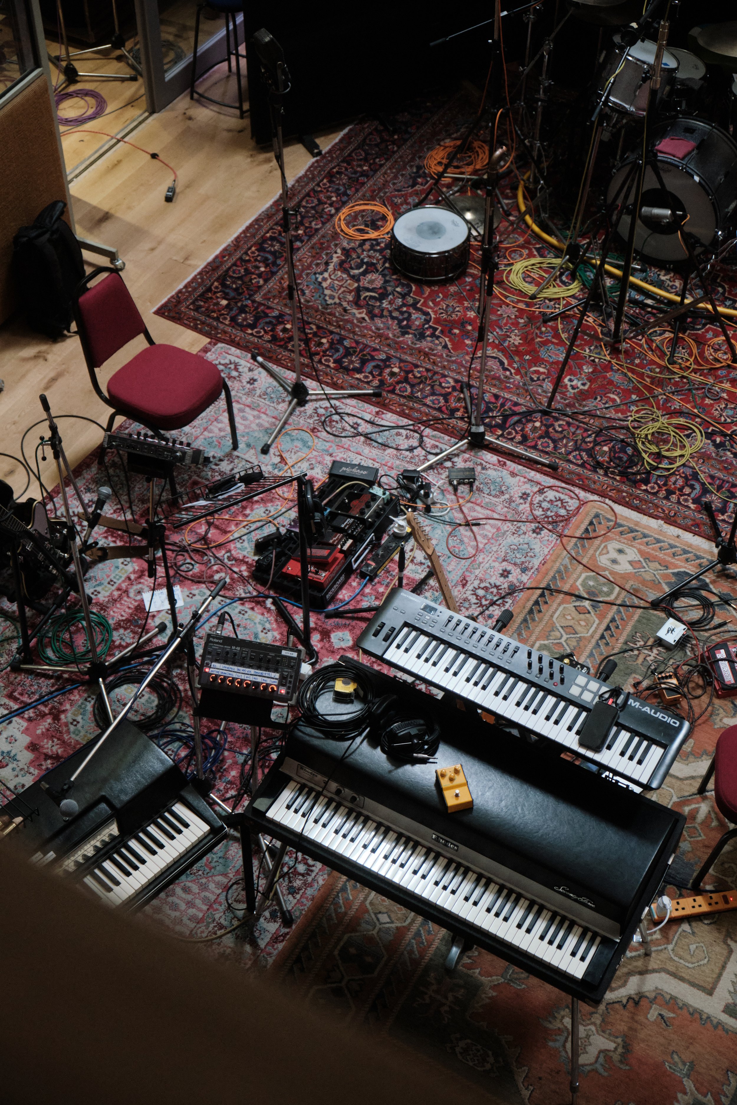 Une configuration de matériel de musique en studio, comprenant un clavier électrique, un synthétiseur, plusieurs microphones, câbles, pédales d'effet, des chaises et un tapis berbère.