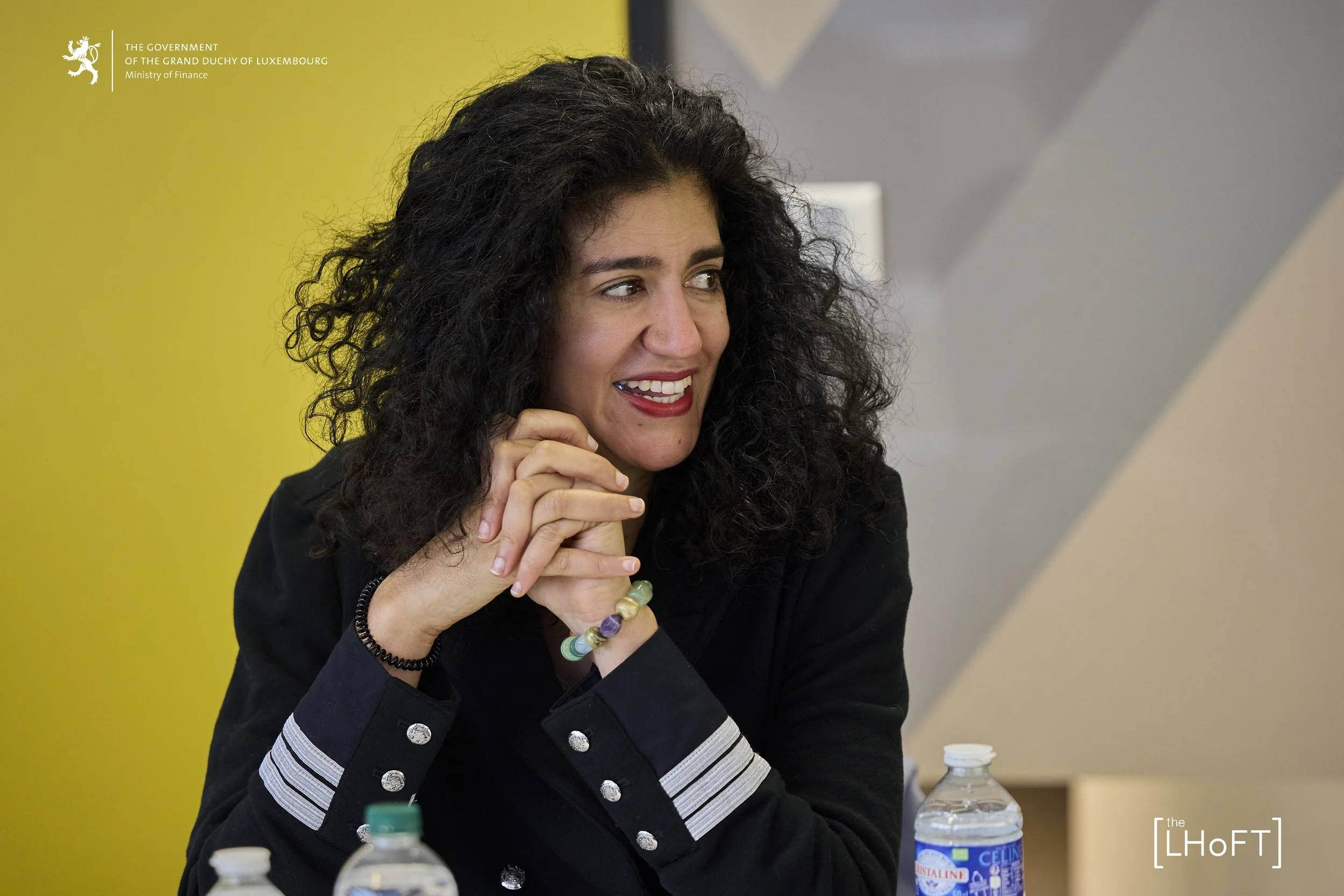 Woman with curly dark hair and a black jacket sitting at a table with water bottles in front of her, smiling and clasping her hands, with a yellow and gray background.
