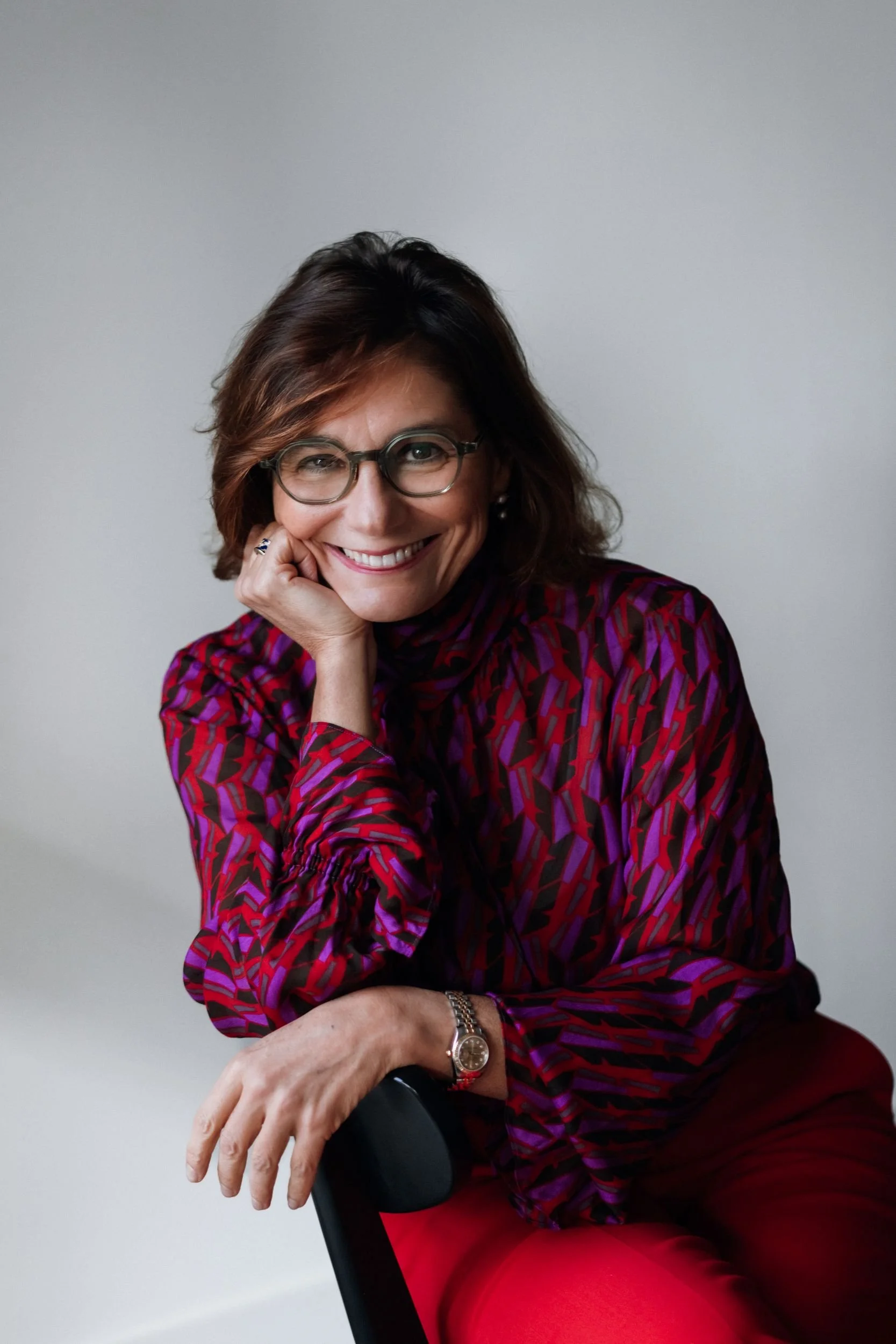 A woman with glasses and shoulder-length brown hair, smiling and resting her chin on her right hand, wearing a red patterned blouse and red pants, sitting on a black chair with a light gray background.
