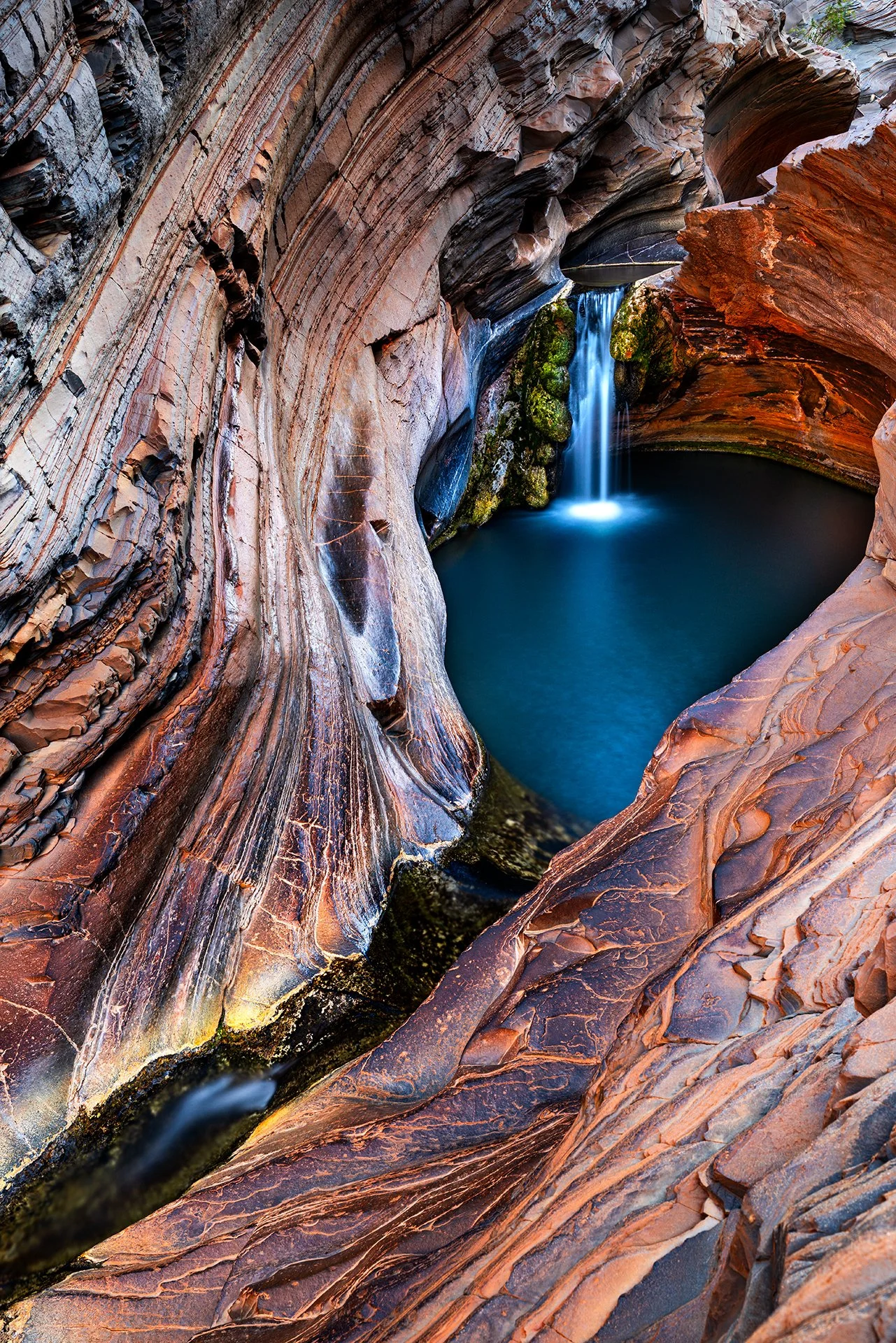 A small waterfall flowing into a pool of water inside a narrow, curved slot canyon with colorful, layered rock walls.