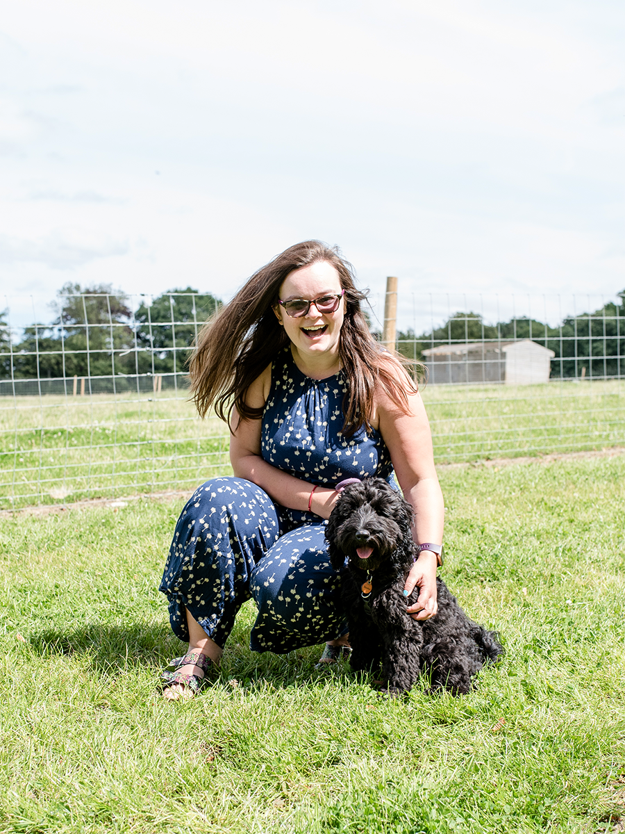 A woman with long brown hair wearing sunglasses and a blue dress with white patterns, crouching on grass, holding a black fluffy dog, outdoors with a wire fence and trees in the background, on a sunny day.