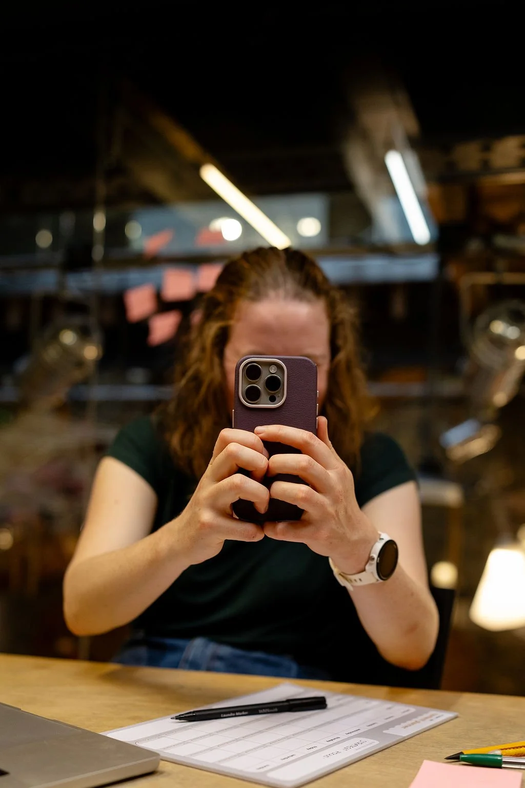 Portrait of brand photographer Beth Hicks taking a photo with a smartphone in a dimly lit setting, with papers and pens on a wooden table in front of her.