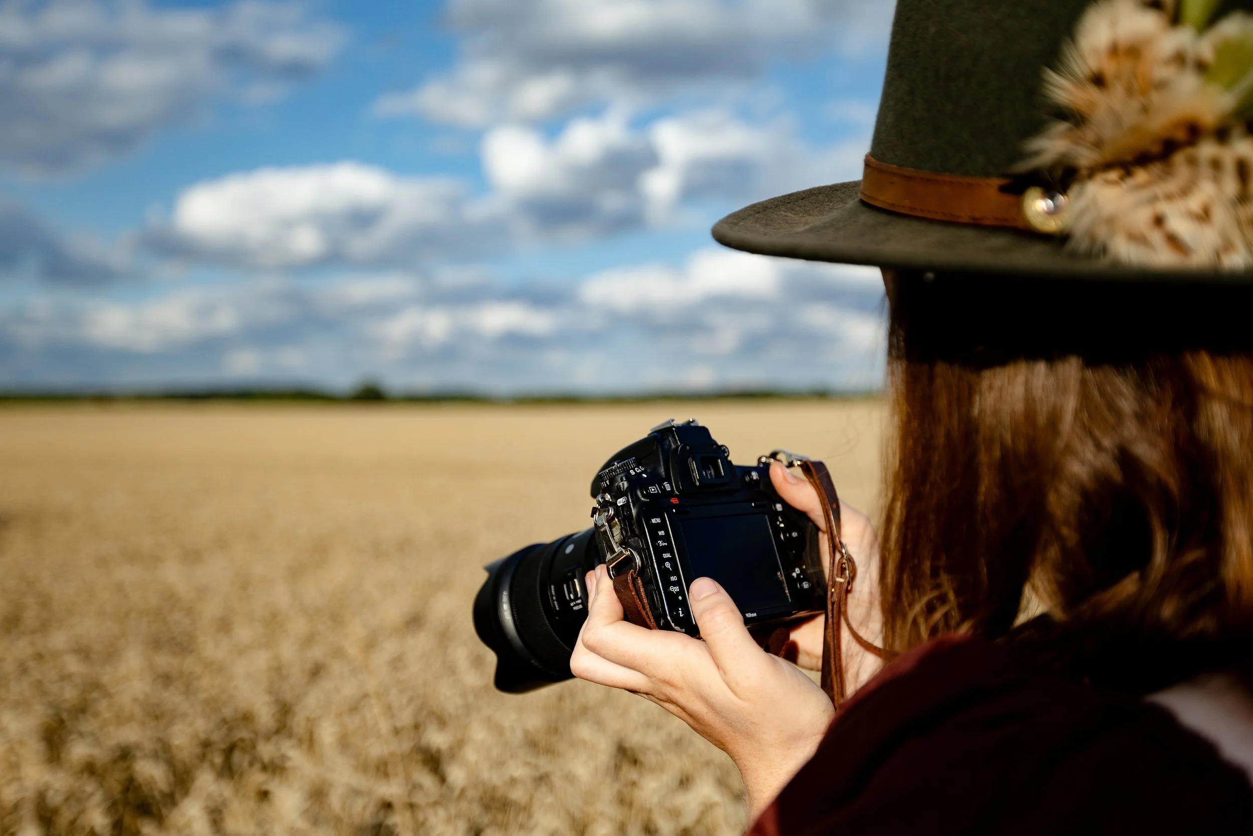 Portrait of brand photographer Beth Hicks with red hair wearing a brown hat with feathers, holding a professional camera, outdoors in a field with a wide blue sky and clouds.