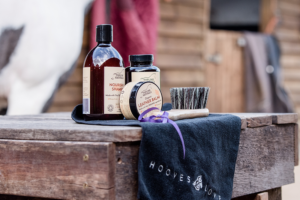 Brand photography of a skincare and grooming products on a wooden surface, including shampoo, leather balm, a small jar, a brush, and a towel with a logo.