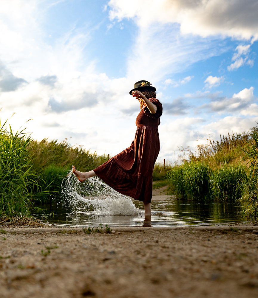 Portrait of brand photographer Beth Hicks wearing a brown dress and black hat with a flower prompt, standing in a small water body, kicking water with her foot, in a grassy natural setting under a partly cloudy sky.