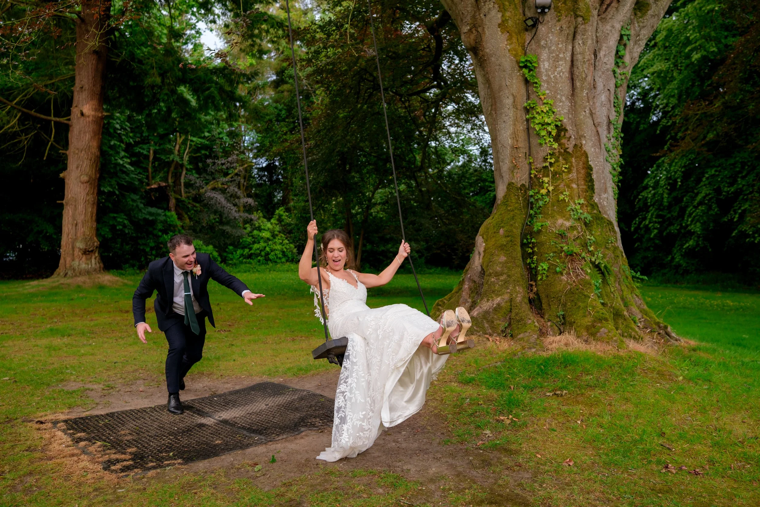 A bride in a white wedding dress sitting on a park swing with a big smile, while a groom in a dark suit and tie pushes her on the swing with joy.