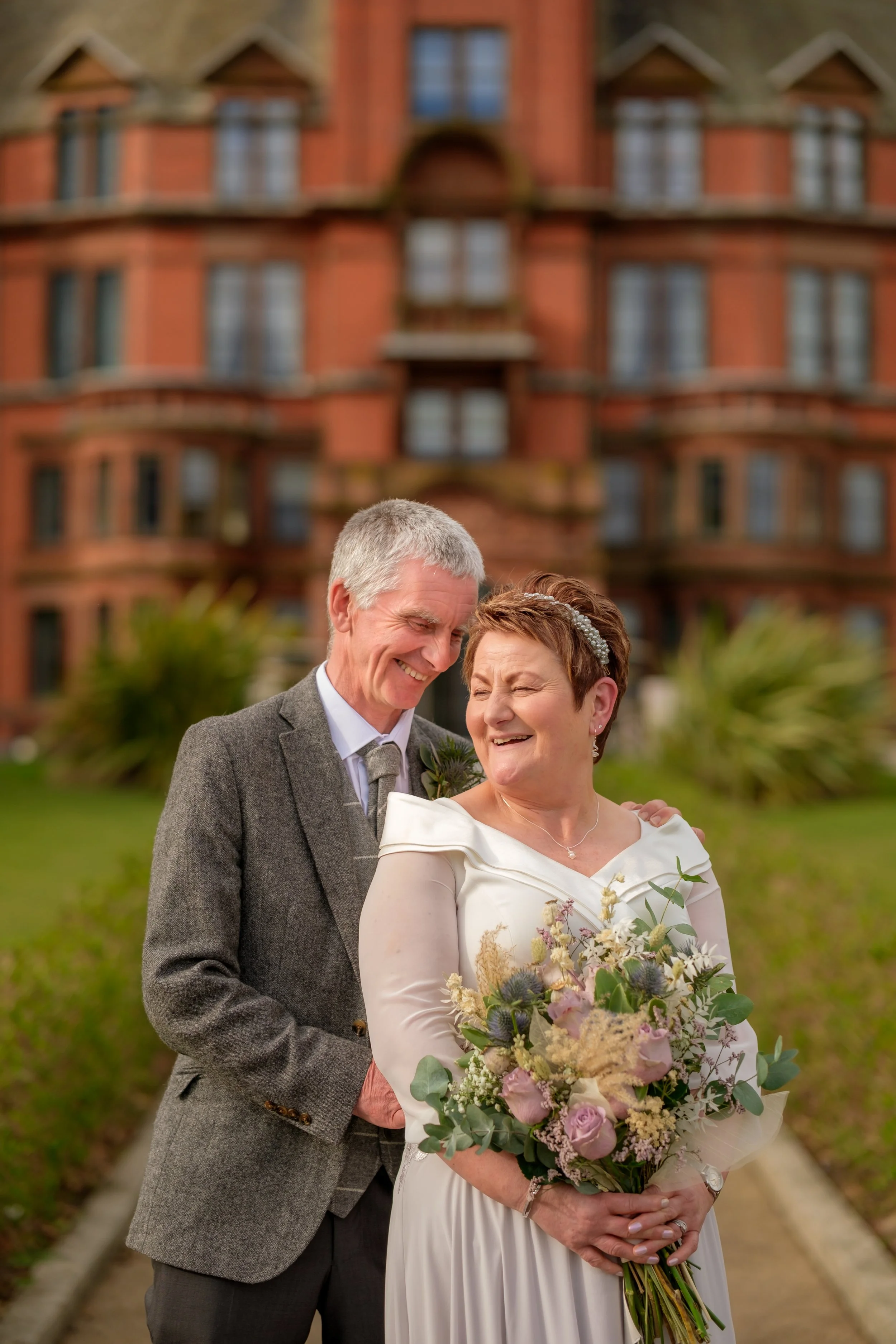 An elderly couple dressed in wedding attire smiling happily outdoors in front of a large brick house, with the woman holding a bouquet of flowers.