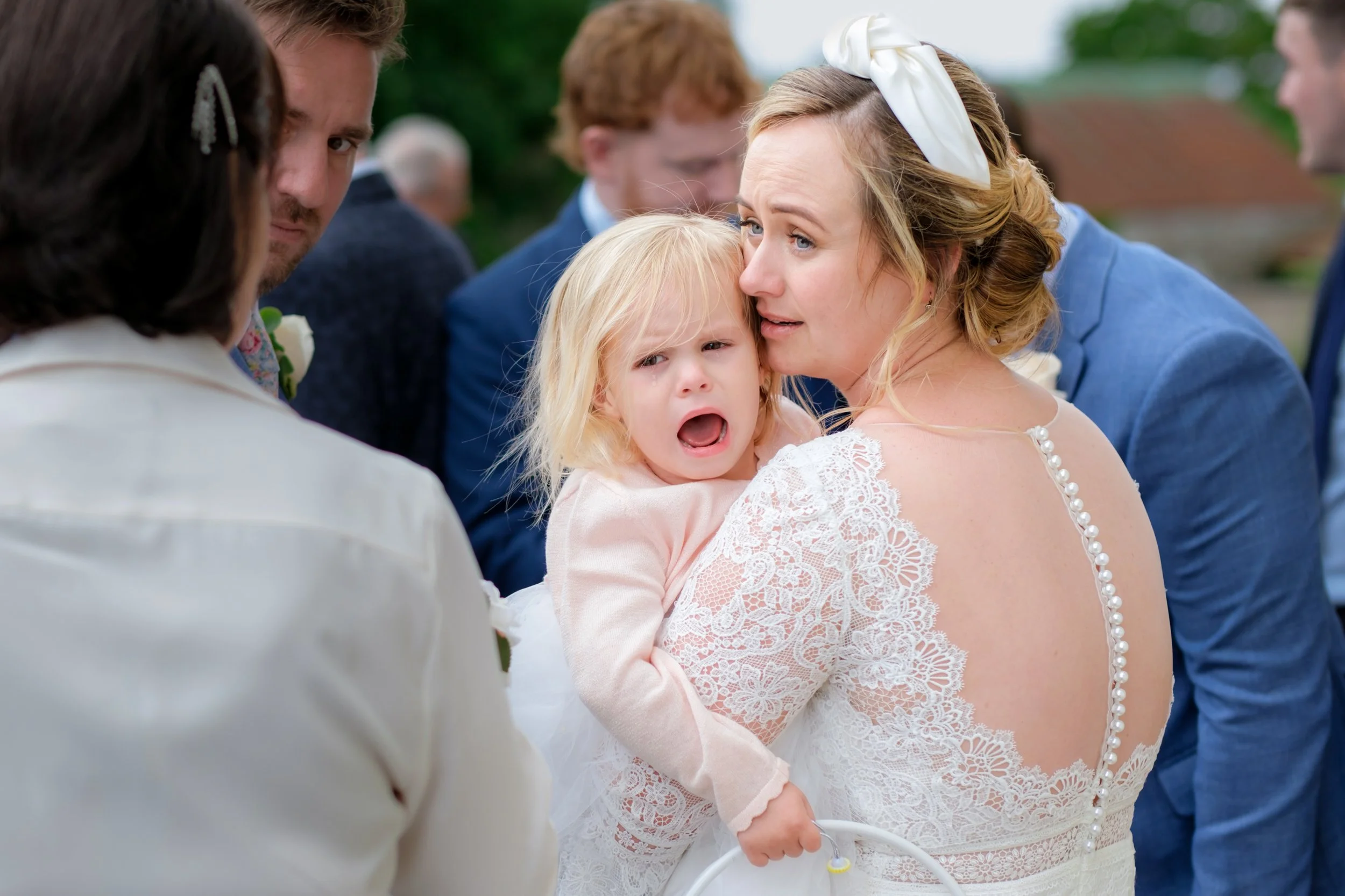 A young girl with blonde hair crying while being held by a woman in a lace dress at an outdoor wedding, with people in the background.
