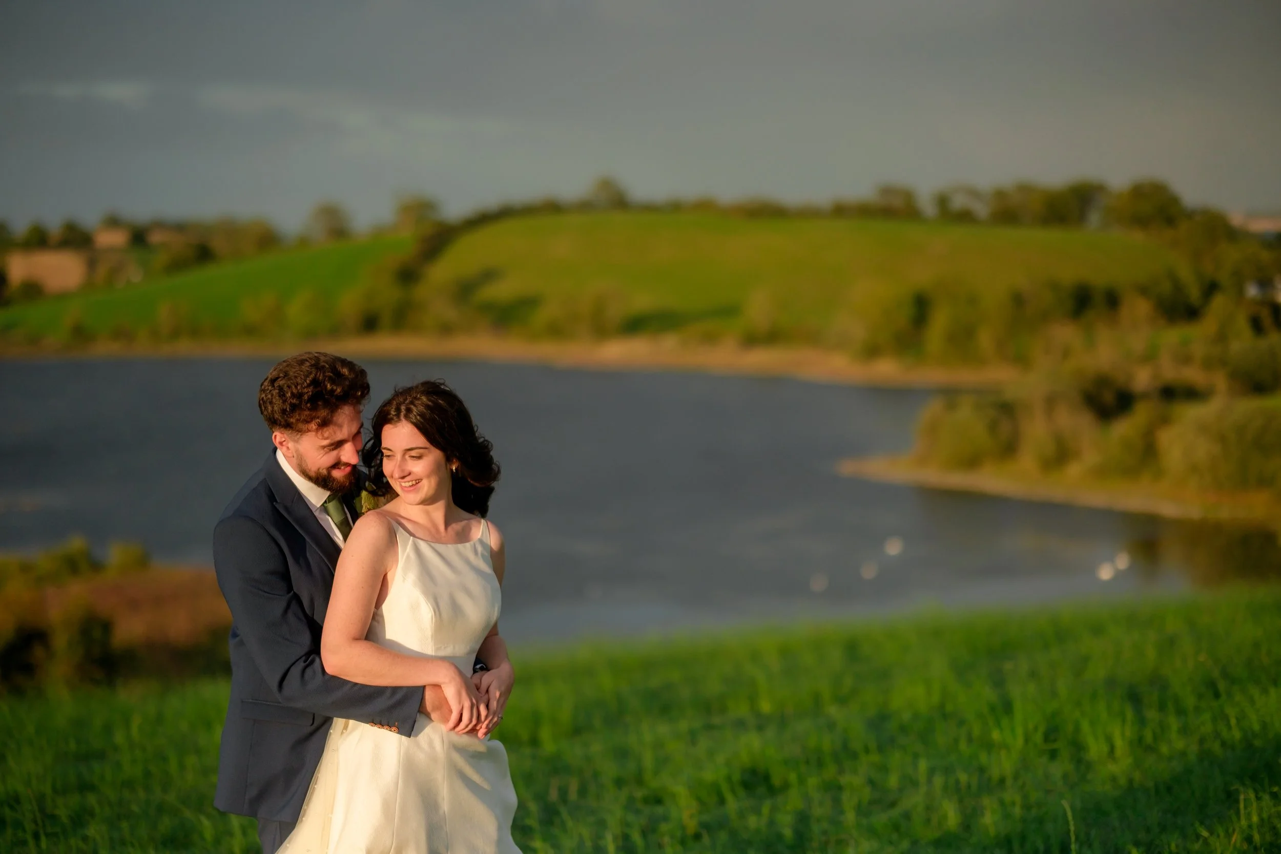 A happy bride and groom standing outdoors by a lake with rolling green hills in the background, smiling and embracing each other during sunset.