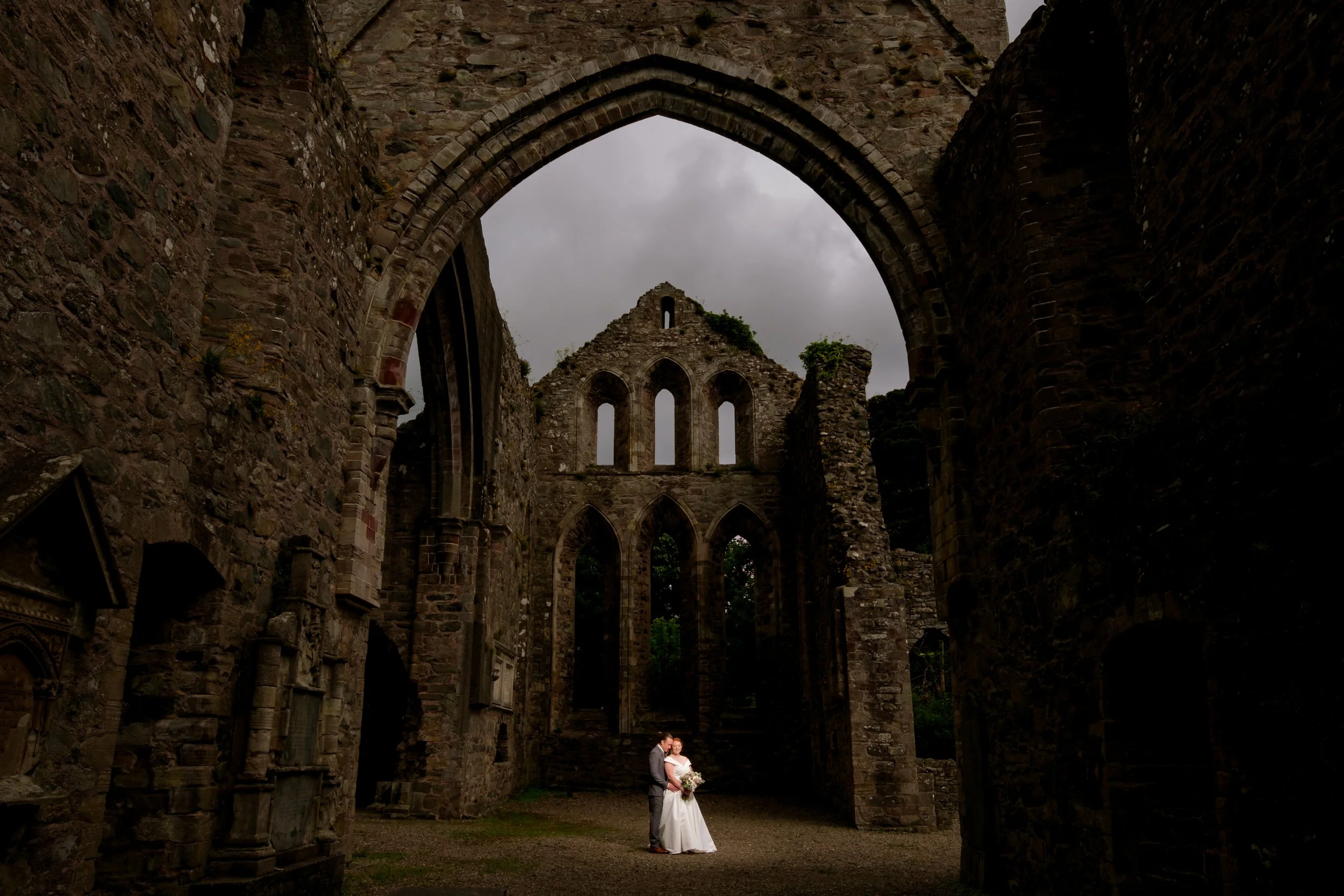 A newlywed couple stands in front of the ruins of an old stone church, embracing each other, with dark clouds overhead.